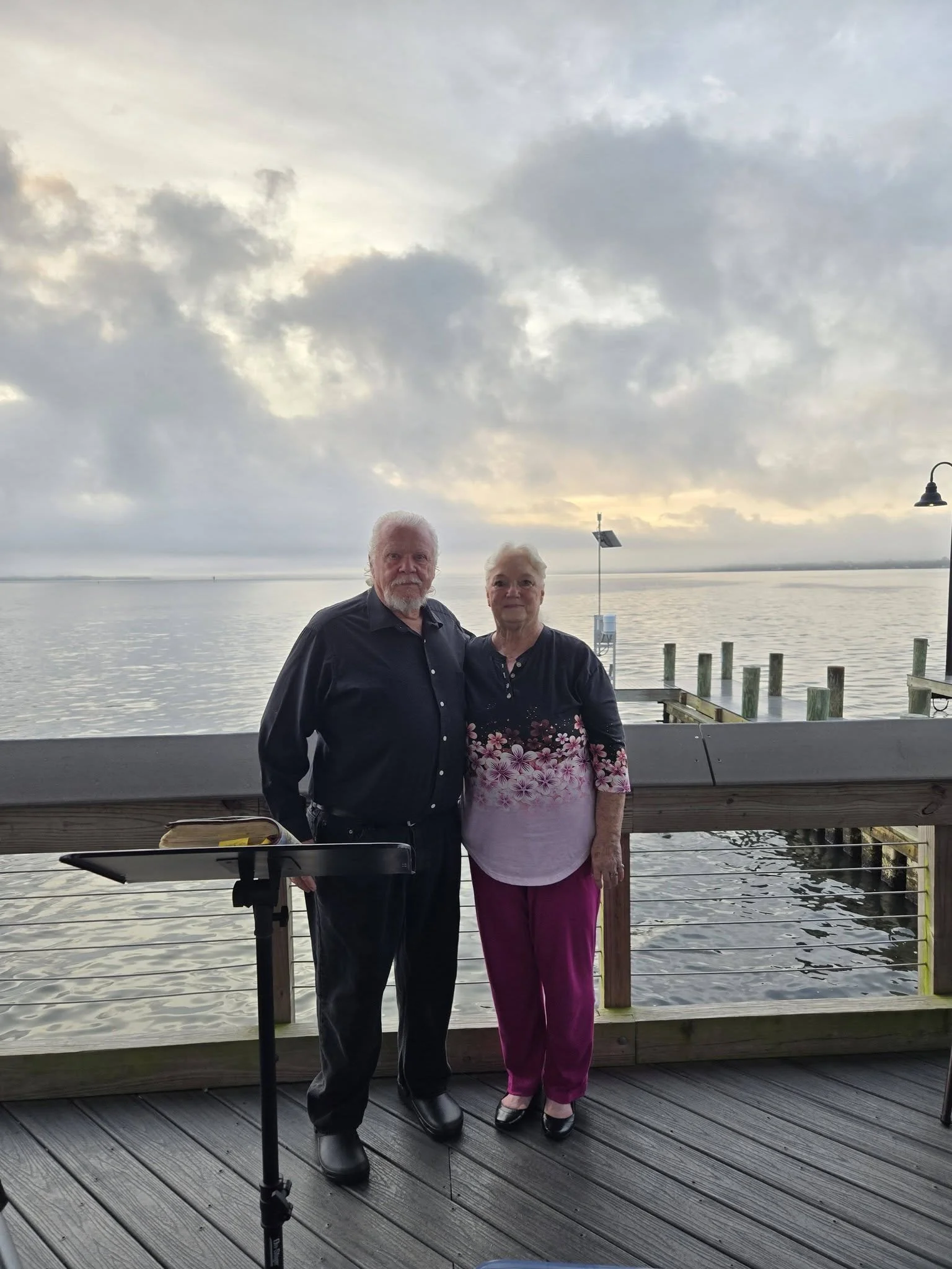 An elderly man and woman standing together on a wooden deck by a body of water, with a cloudy sky overhead, during sunset or sunrise.