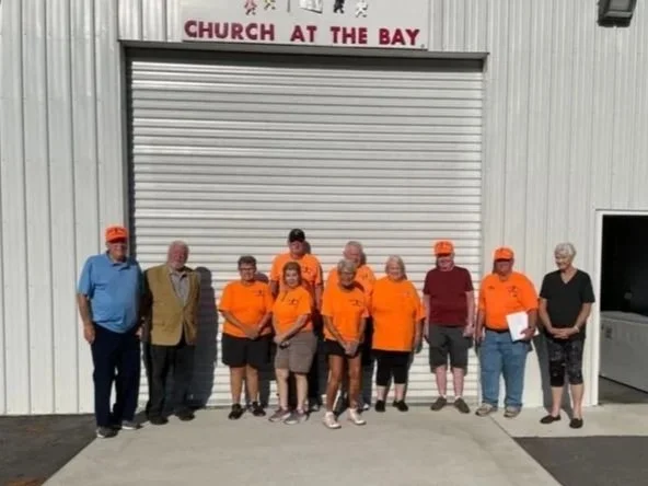 Group of ten people standing in front of a white building with a closed garage door and a sign that says 'CHURCH AT THE BAY'.