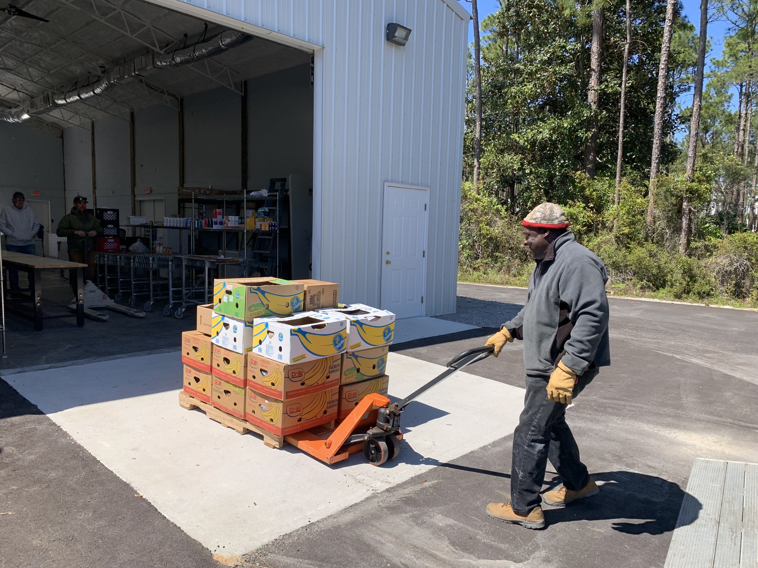Man operating a pallet jack with boxes of bananas outside a warehouse with trees in the background.