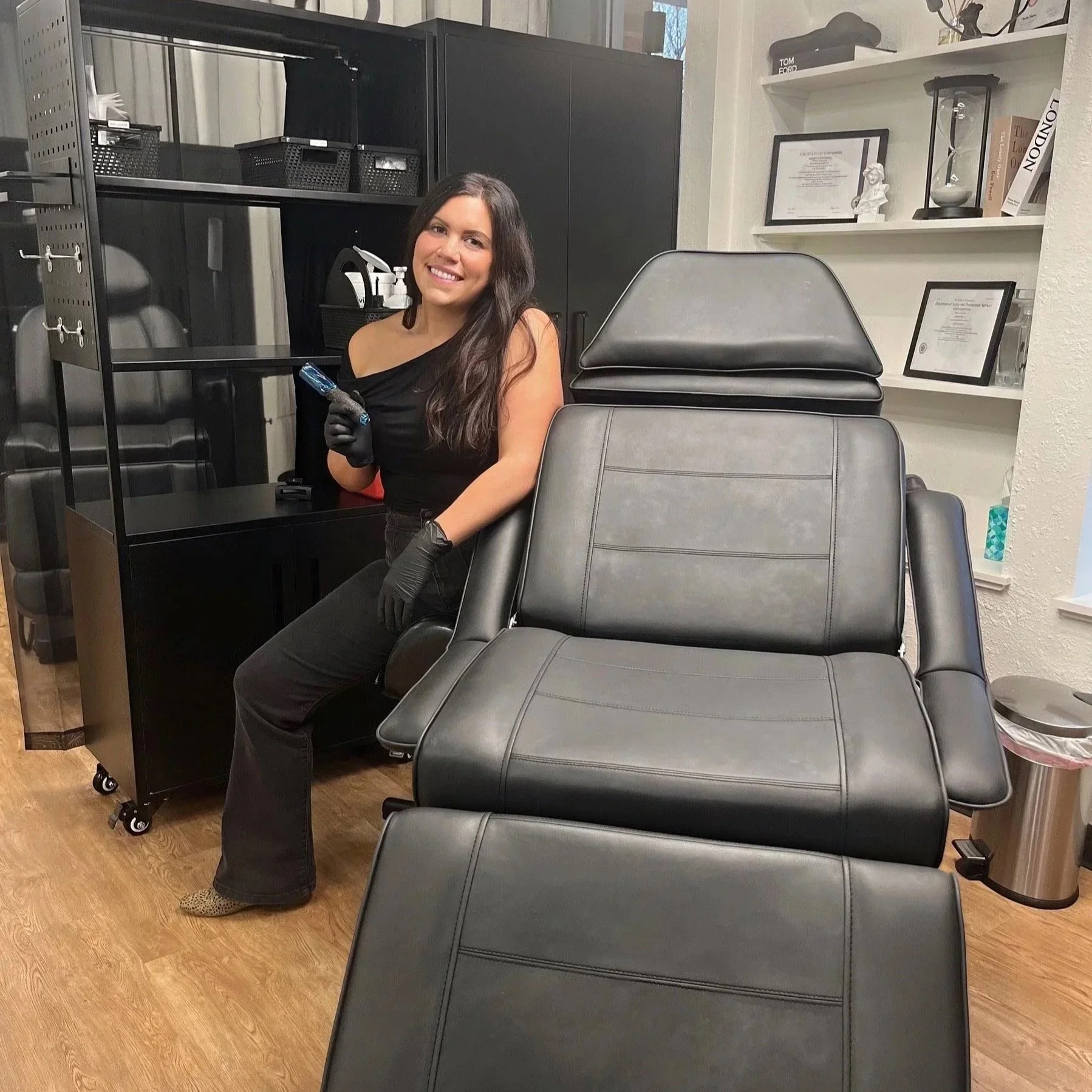 A woman with long brown hair wearing a black off-shoulder top, black gloves, and black pants standing in a room with black furniture, holding a tattoo machine, smiling at the camera.