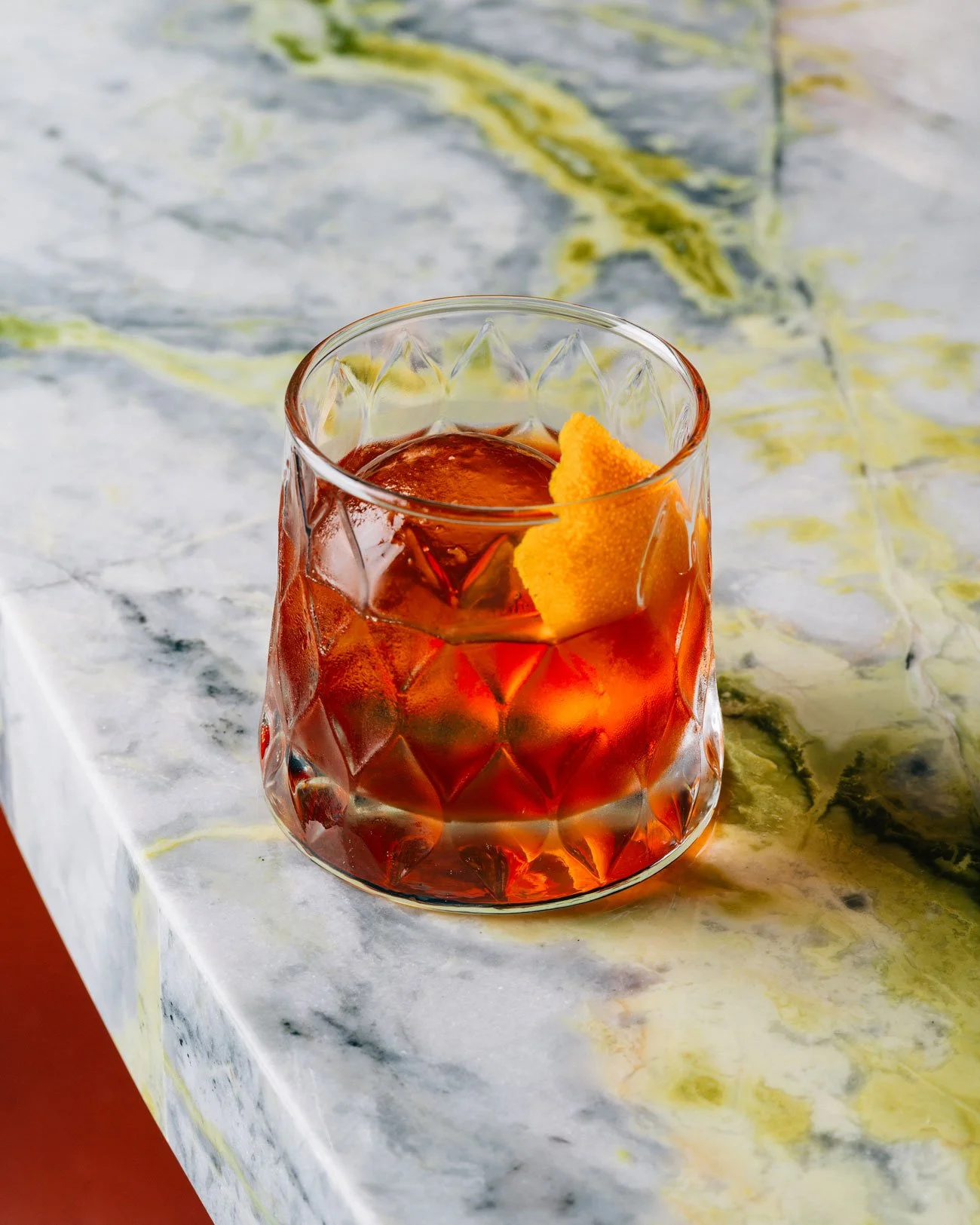 A glass with a textured pattern containing a Negroni cocktail garnished with a lemon twist, placed on a marble surface.