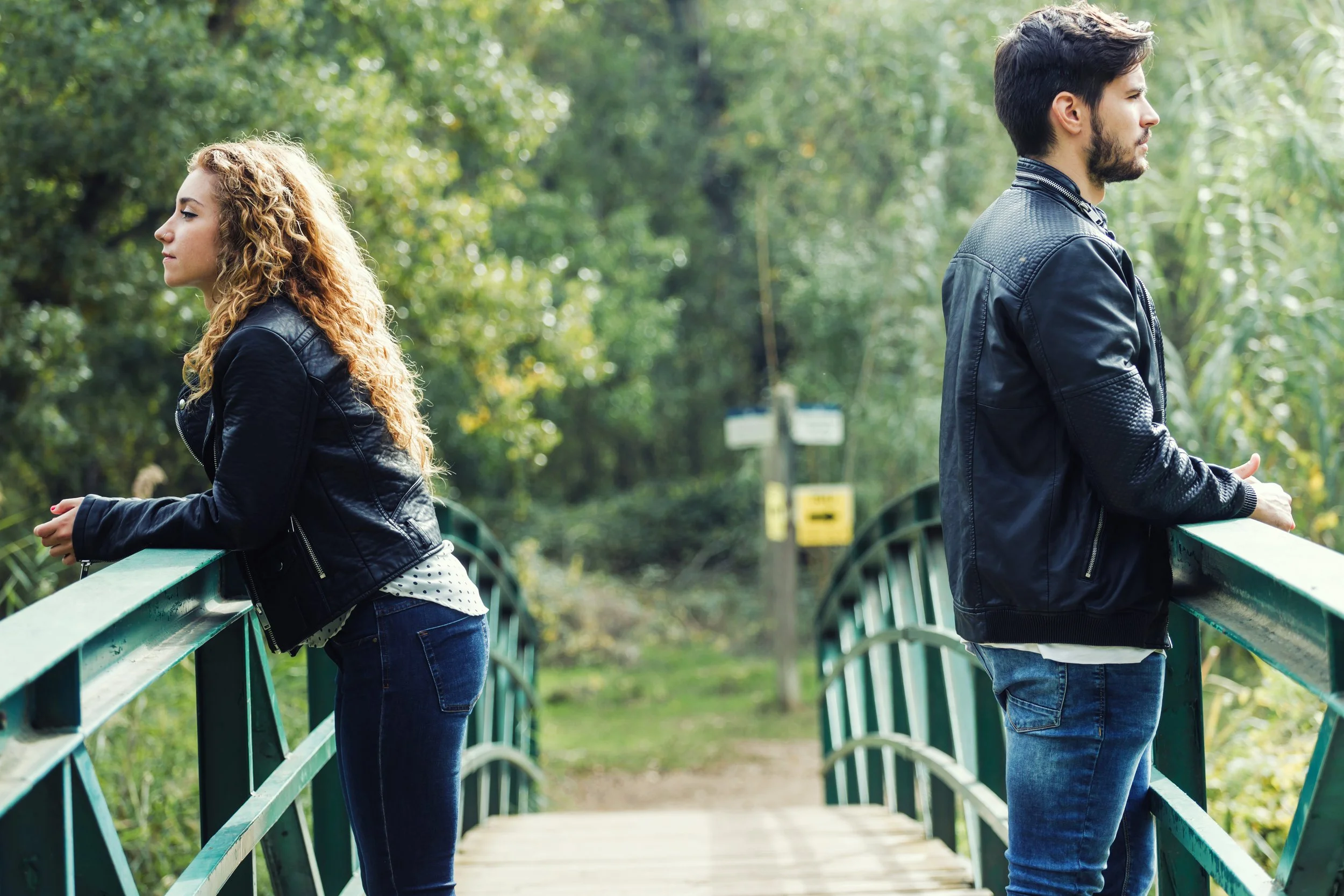 A young couple stands on a bridge facing opposite directions while navigating challenge in their relationship.