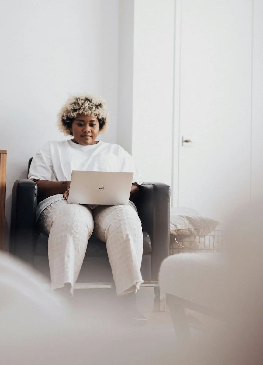 A woman wearing white sits in a comfortable chair while logging onto her laptop for virtual therapy.