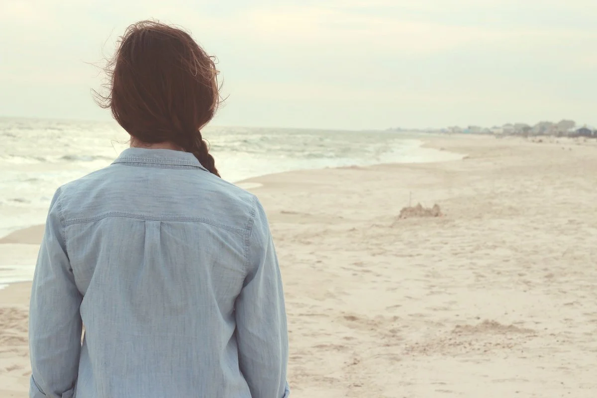 A woman with long dark hair looks to the horizon while standing on the beach.