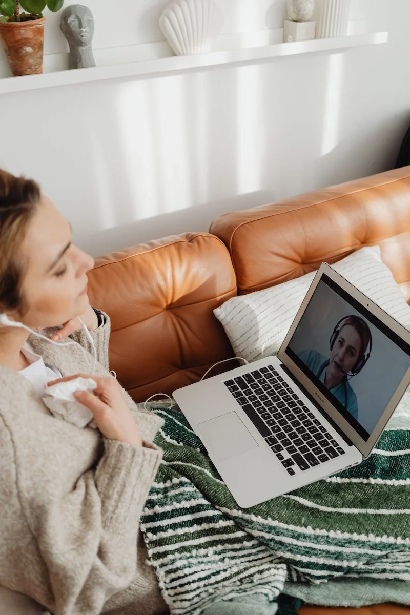 A woman smiles and waves at her laptop camera during a virtual therapy session.