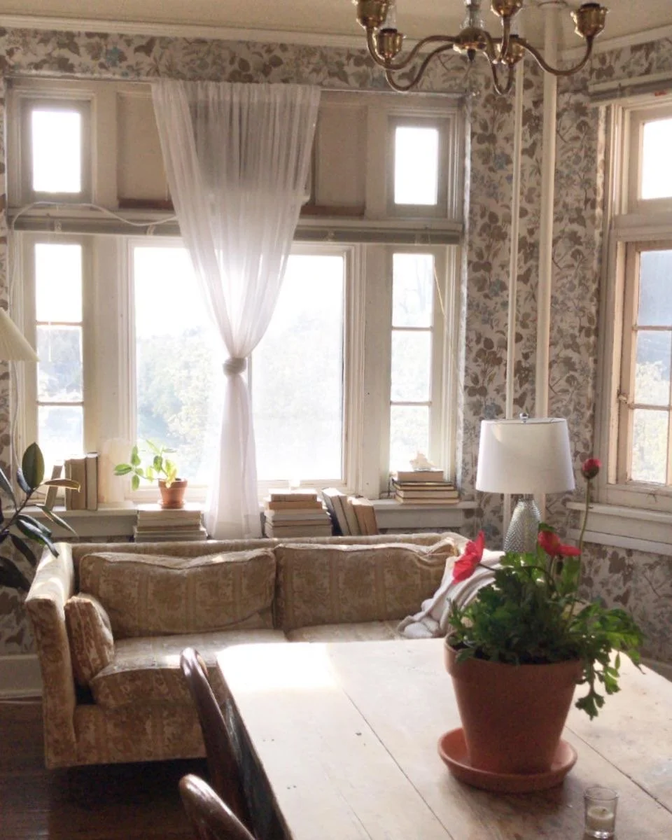 Cozy living room with large windows, floral wallpaper, a vintage sofa, wooden table with potted plant and flowers, closet books, and a chandelier.