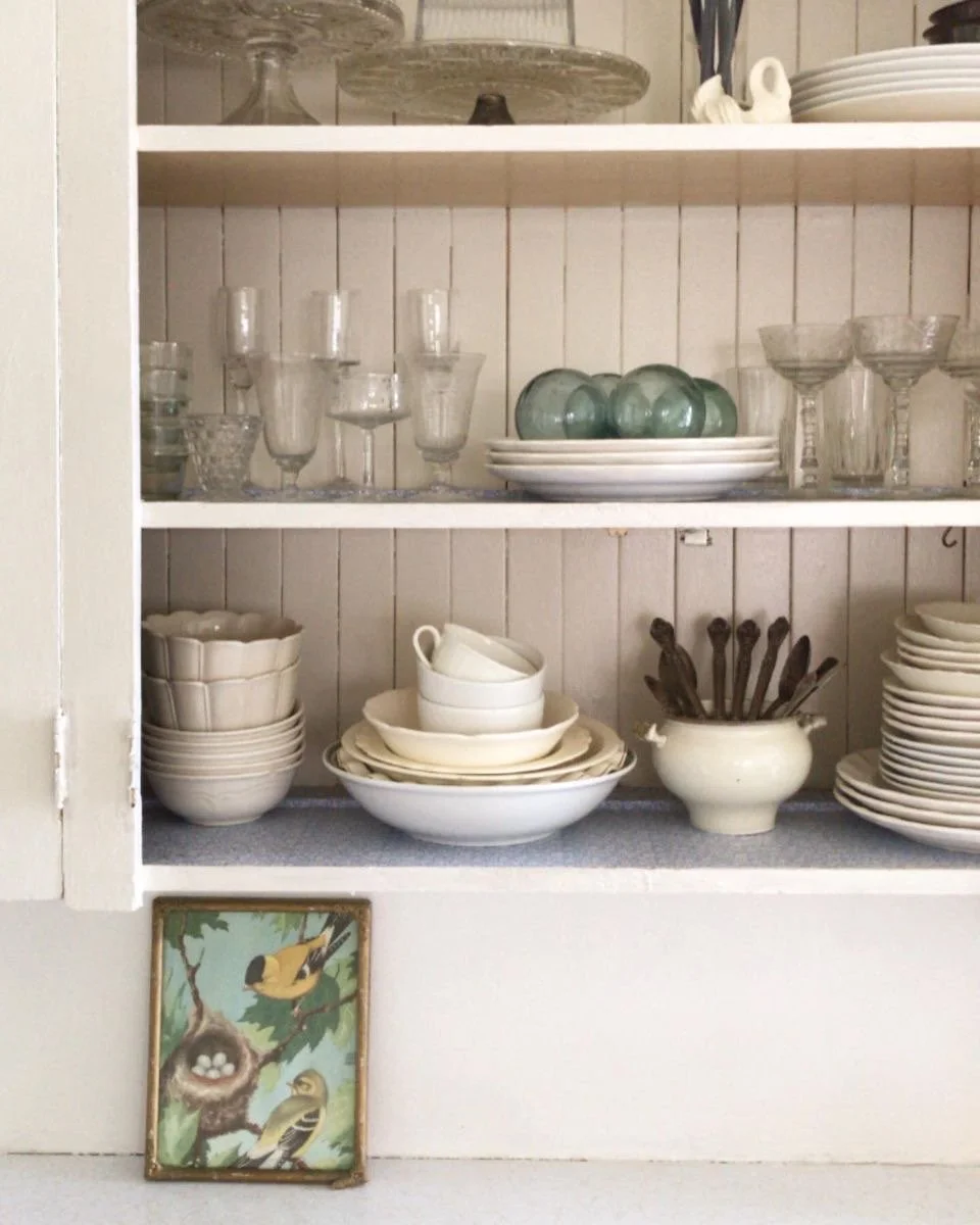 A white kitchen cabinet with glassware, dishes, and utensils inside. There are stacks of bowls, plates, and cups, along with a small painting of birds near the bottom on the wall.