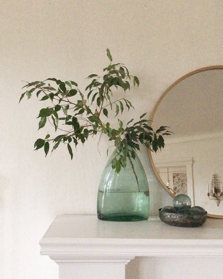 A green glass vase with leafy branches on a white mantel, with a round mirror and decorative stones nearby.