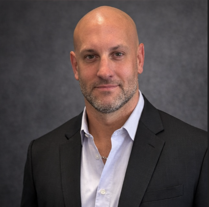 Professional headshot of a bald man with a beard, wearing a black suit jacket and white dress shirt, against a dark gray background.