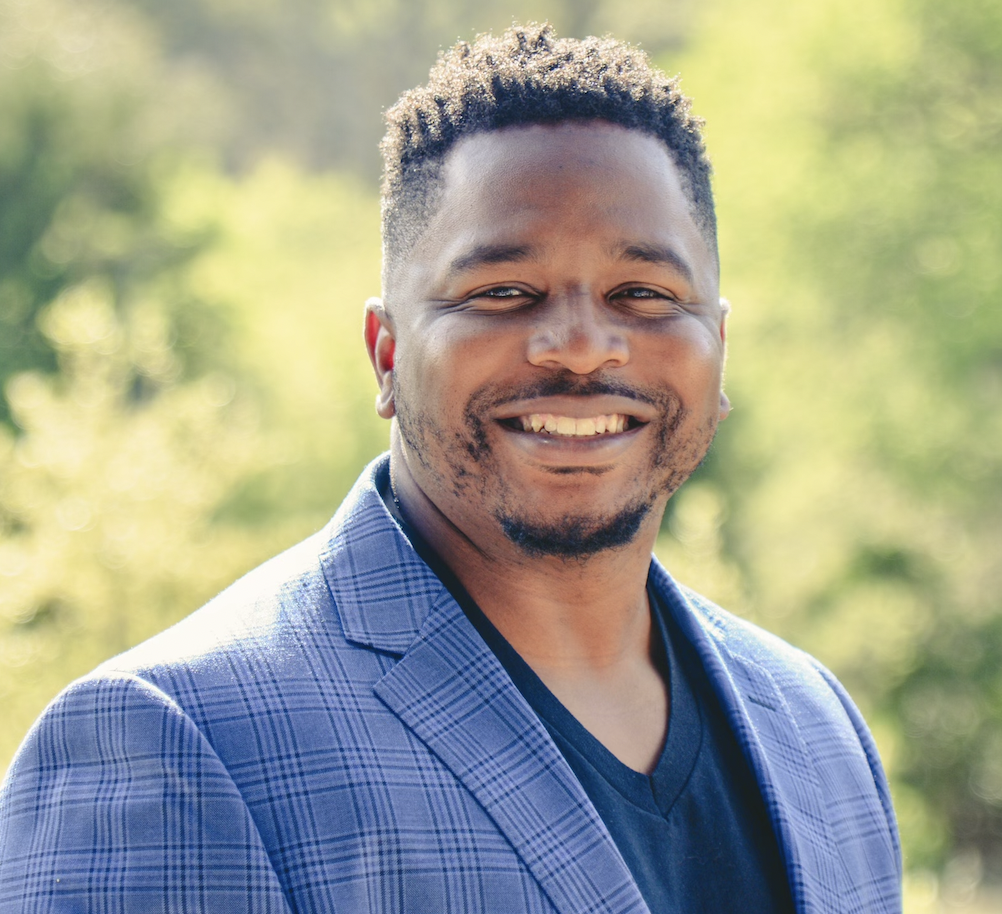 Smile of a man outdoors with blurred green trees in the background, wearing a blue plaid blazer and black shirt.