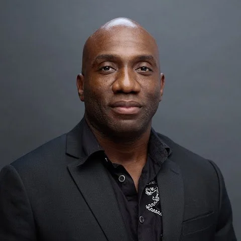 Professional headshot of a bald man in a black suit and black shirt against a dark gray background.