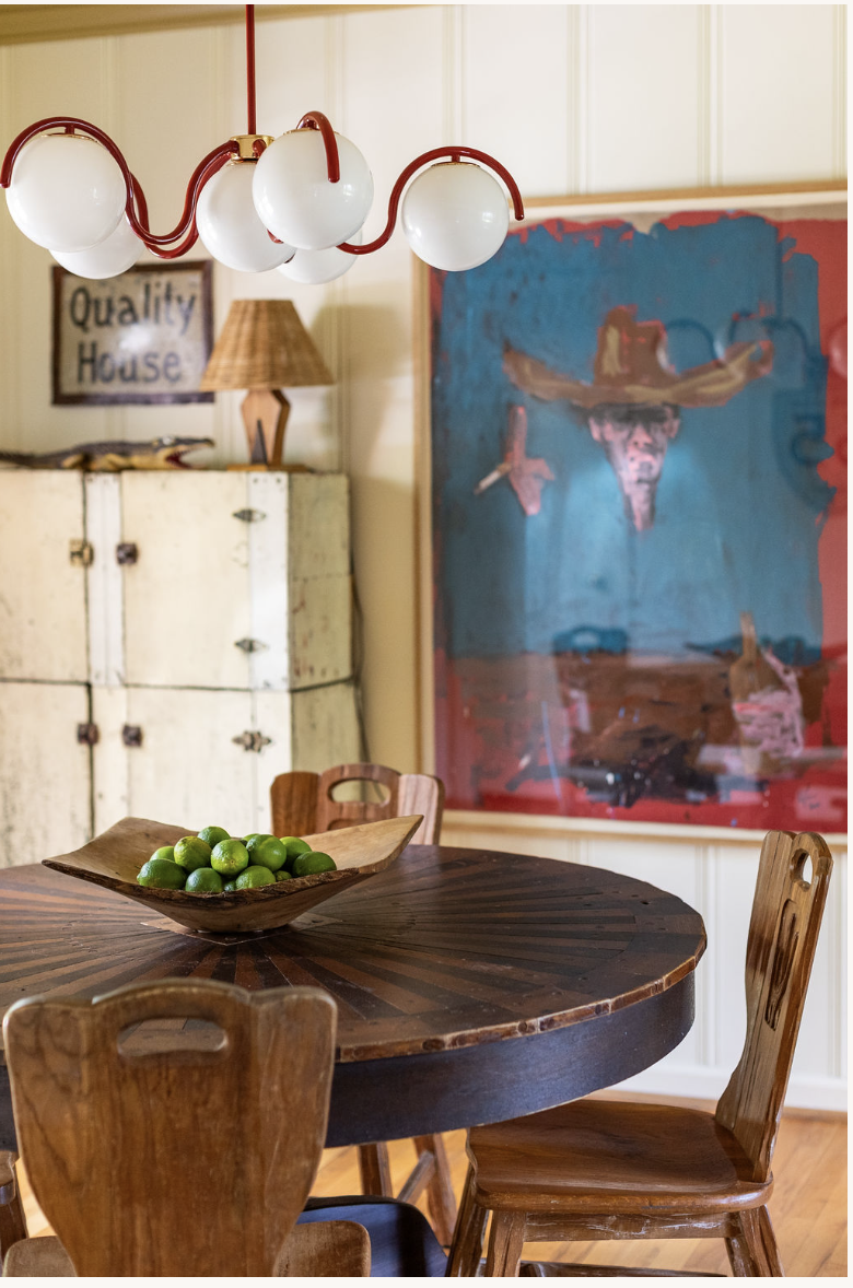 A wooden dining table with a bowl of green limes, surrounded by wooden chairs, in a room with artwork and decorative items on the walls.