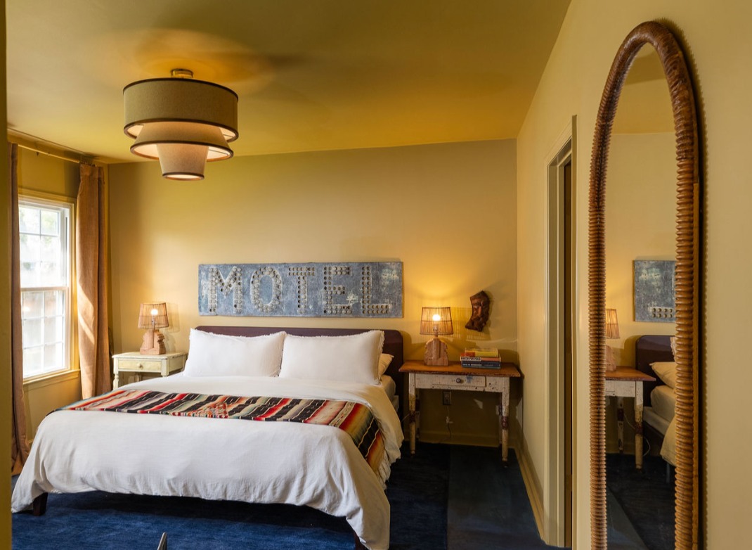 A cozy bedroom with a large bed, two bedside lamps, a 'MOTEL' sign above the bed, a mirror on the right, and a window with curtains letting in natural light.