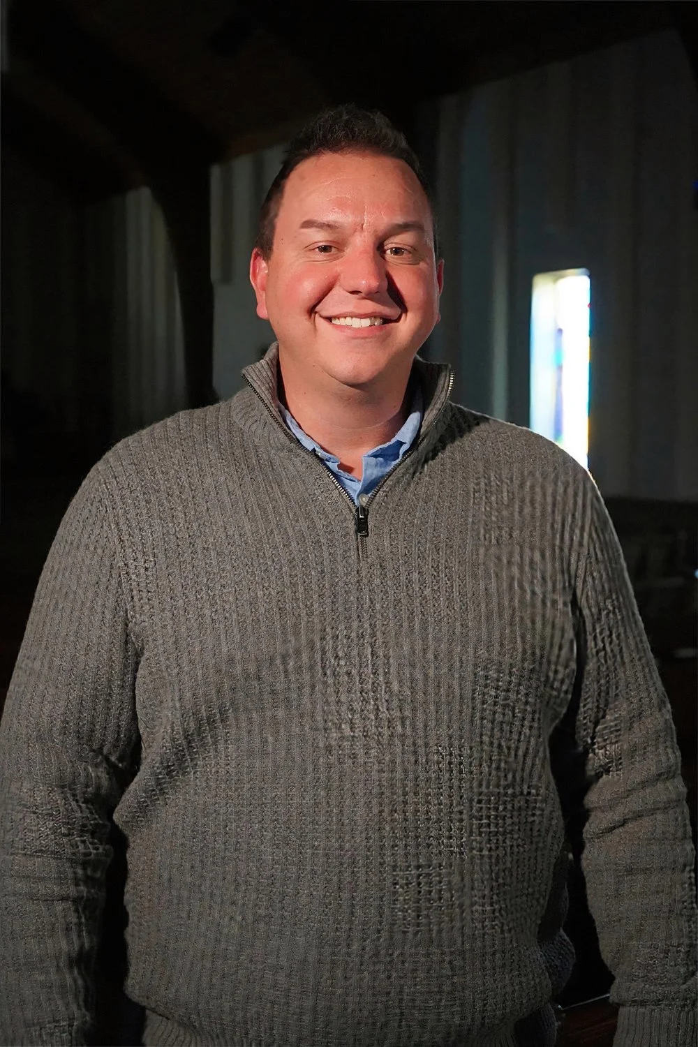 A smiling man with short dark hair in a gray sweater stands inside a dimly lit room with vertical curtains and stained glass window in the background.