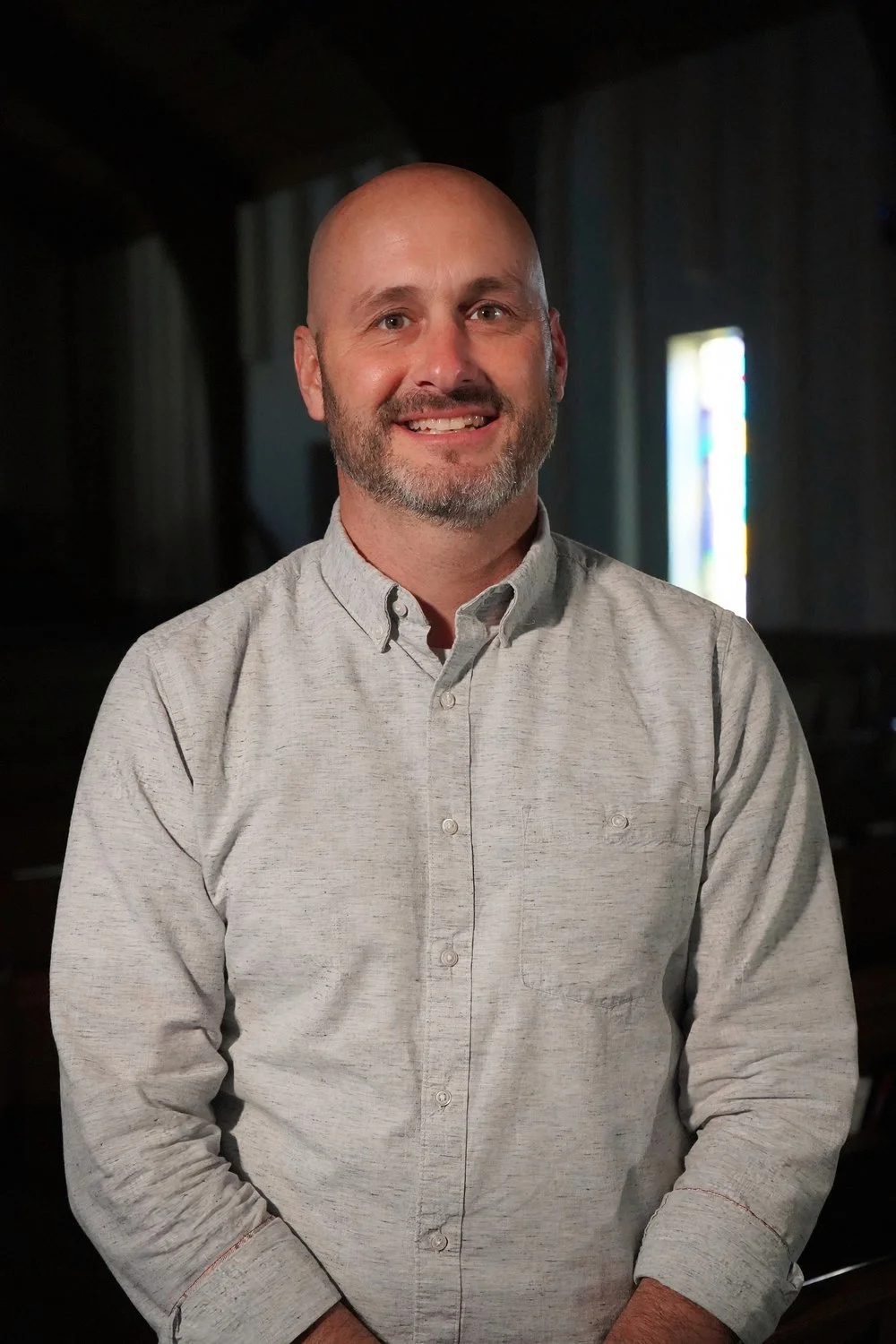 A man with a shaved head and beard, wearing a light gray button-up shirt, standing indoors with a blurred background featuring stained glass and dark walls.