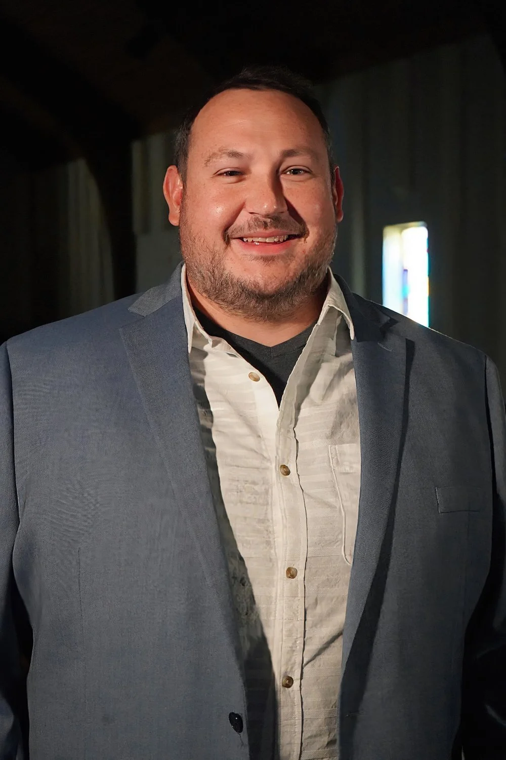 A smiling man in a gray suit and white shirt standing indoors with dim lighting and a stained glass window in the background.