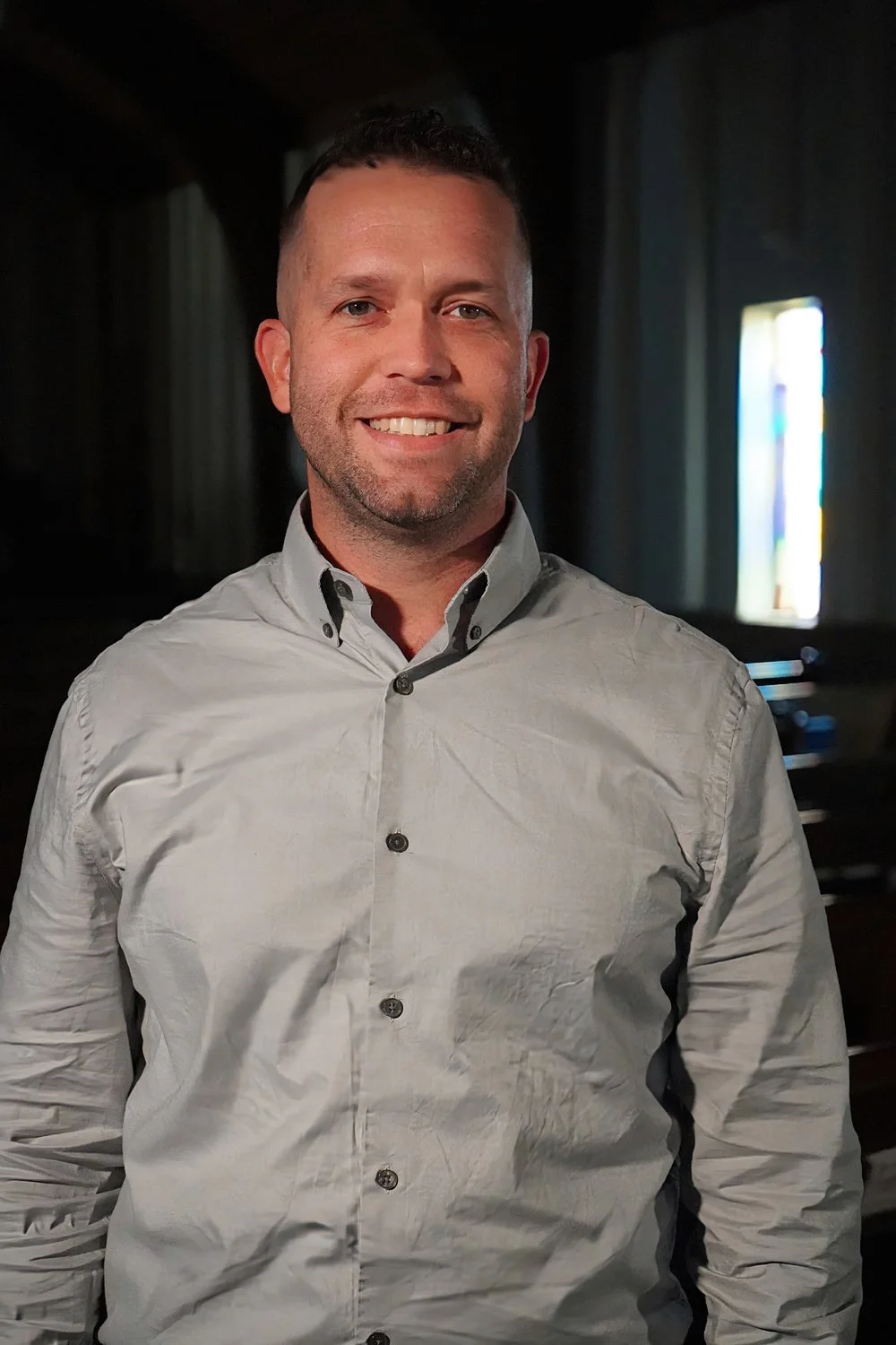 A smiling man with short dark hair and a beard, wearing a light gray button-down shirt, standing inside a dimly lit church with stained glass windows in the background.