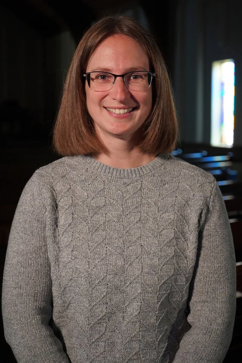 A woman with shoulder-length brown hair, wearing glasses and a gray cable-knit sweater, smiling at the camera in a dimly lit room with a stained glass window in the background.