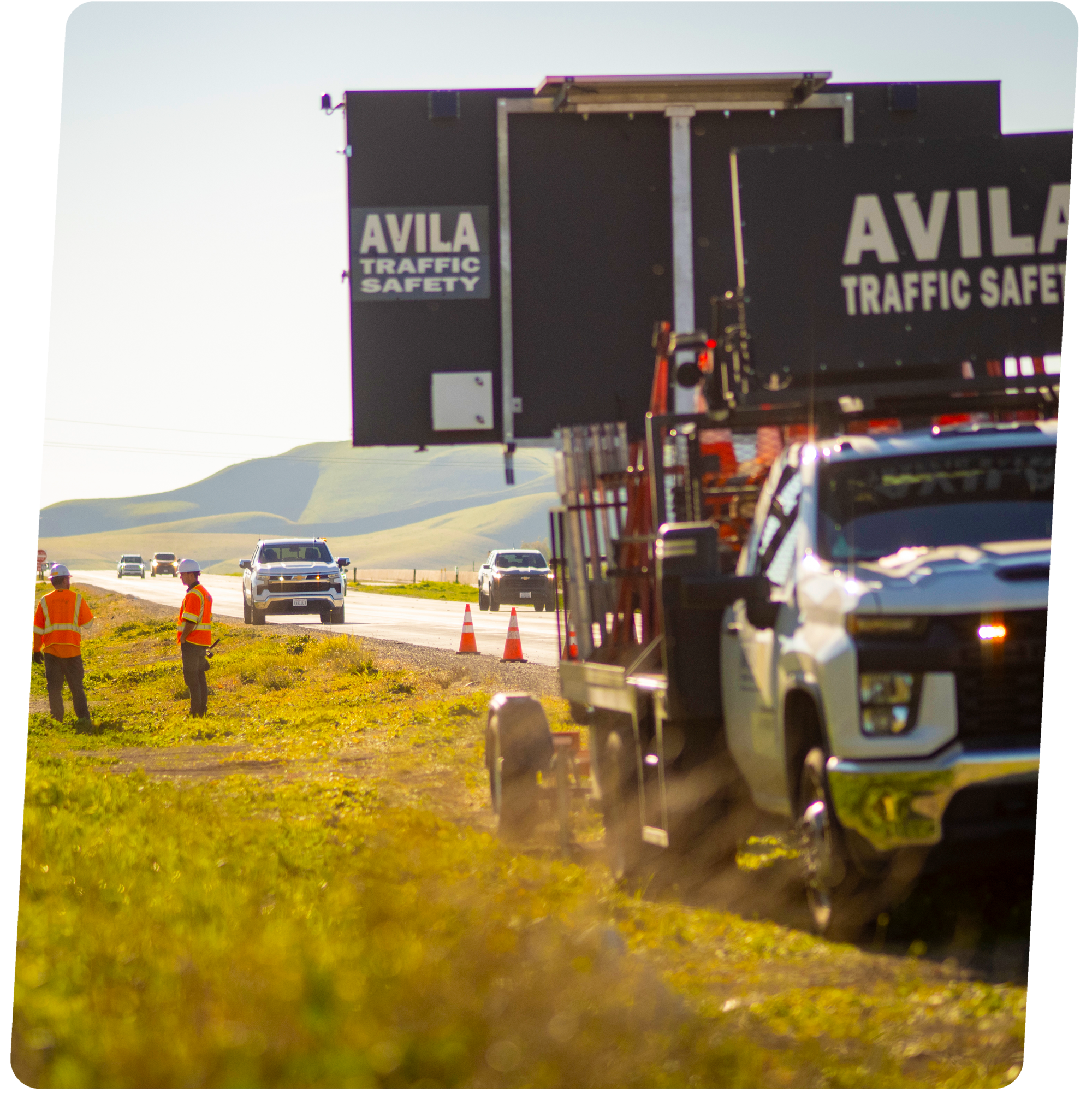 Construction workers on the roadside near traffic cones and a large black sign on a truck, with cars traveling on a highway in a rural landscape with hills in the background.