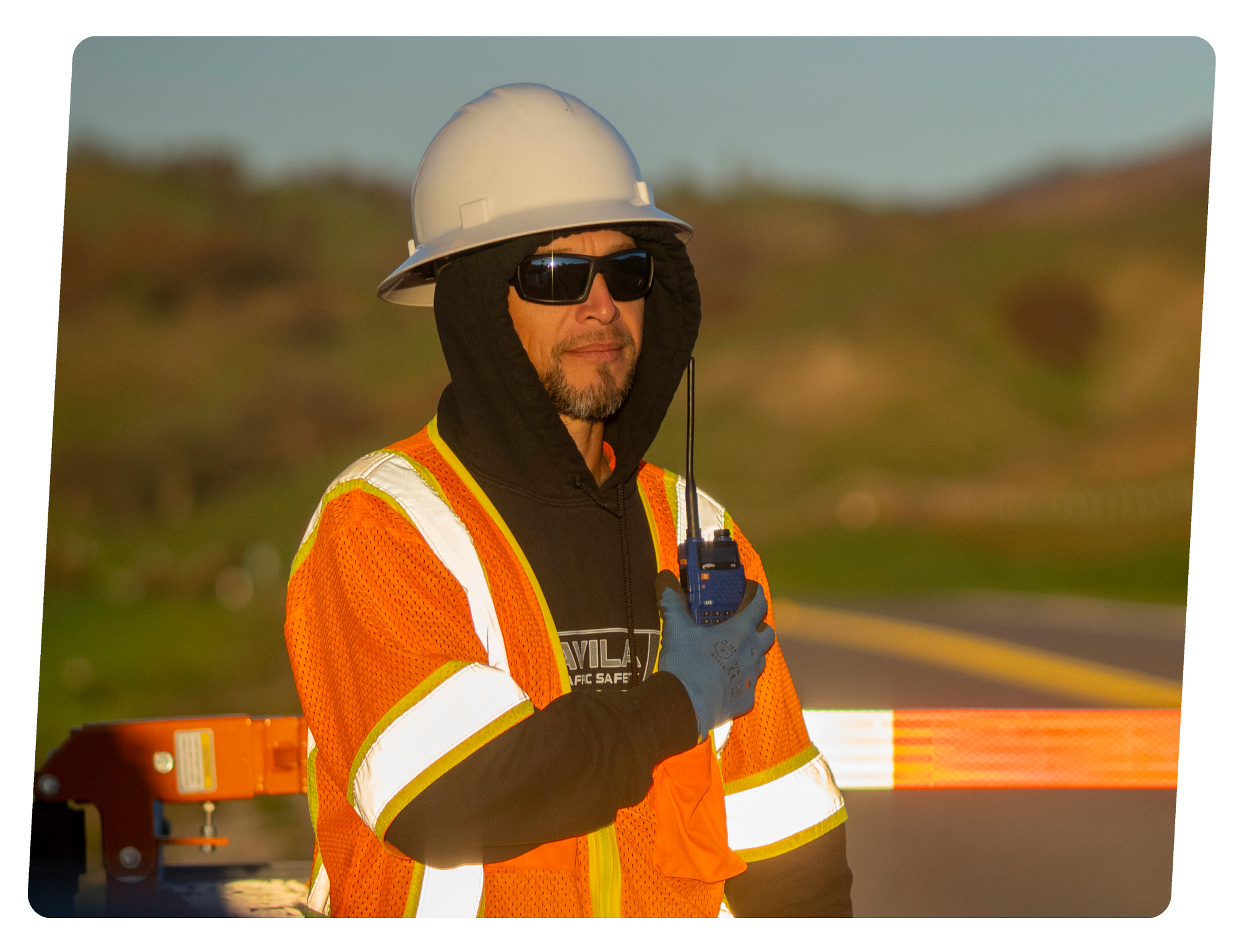 A male traffic control worker wearing a white hard hat, sunglasses, an orange reflective safety vest, black hoodie, and blue gloves. He holds a portable radio in his right hand. The background shows a blurred landscape with hills.