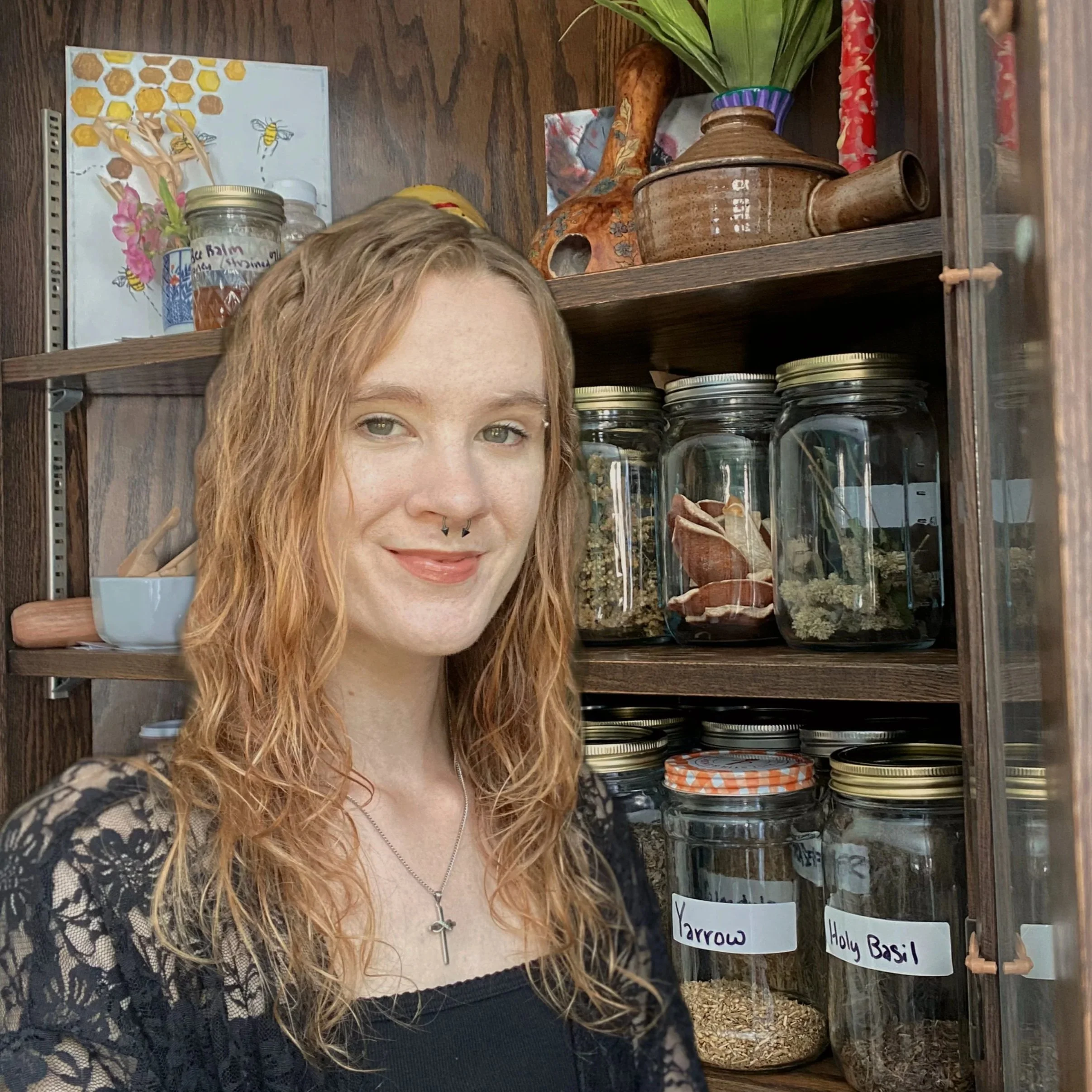 A woman with curly red hair, light skin, and a septum piercing smiling in front of a wooden pantry shelf filled with jars of herbs, spices, and dried ingredients. She wears a black lace top and a necklace with a cross pendant.