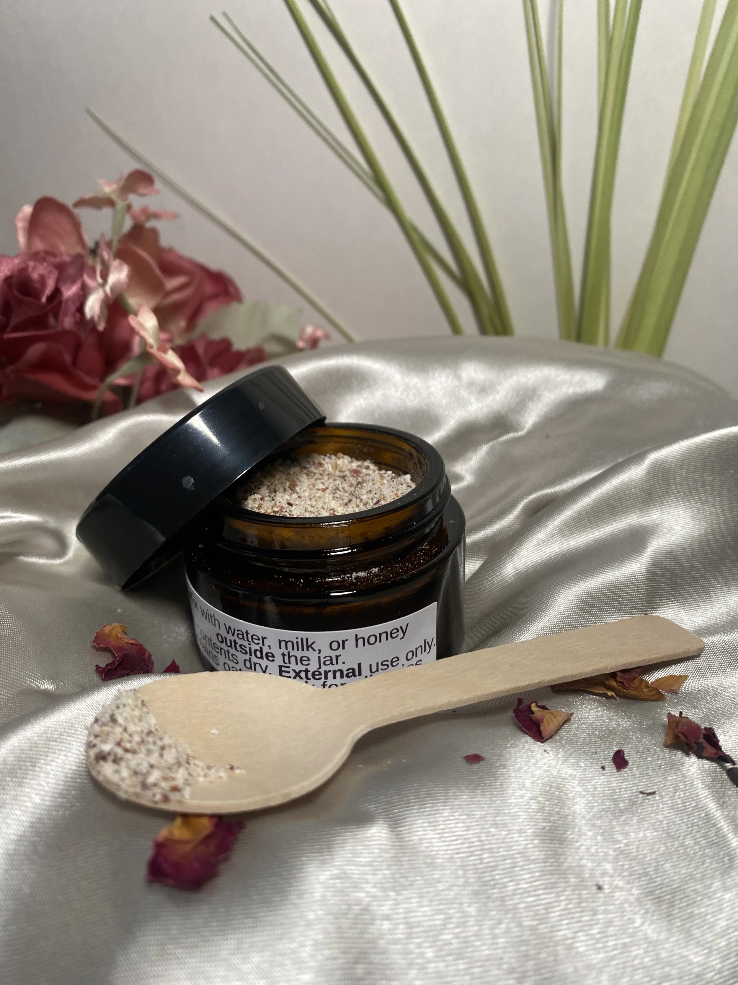 An open dark brown jar with a white label containing instructions, sitting on a satin fabric surface, with a small wooden spoon filled with a granular substance, surrounded by dried rose petals and green leaves in the background.