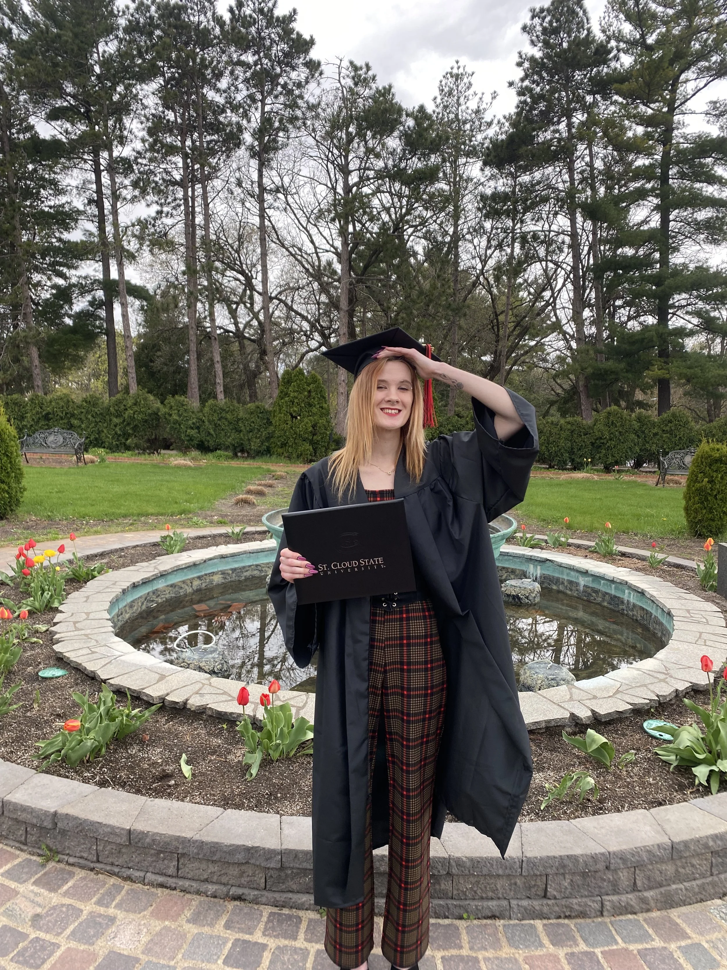 A young woman in a graduation cap and gown standing in front of a small fountain, holding a diploma from St. Cloud State University, smiling with her hand on her forehead.