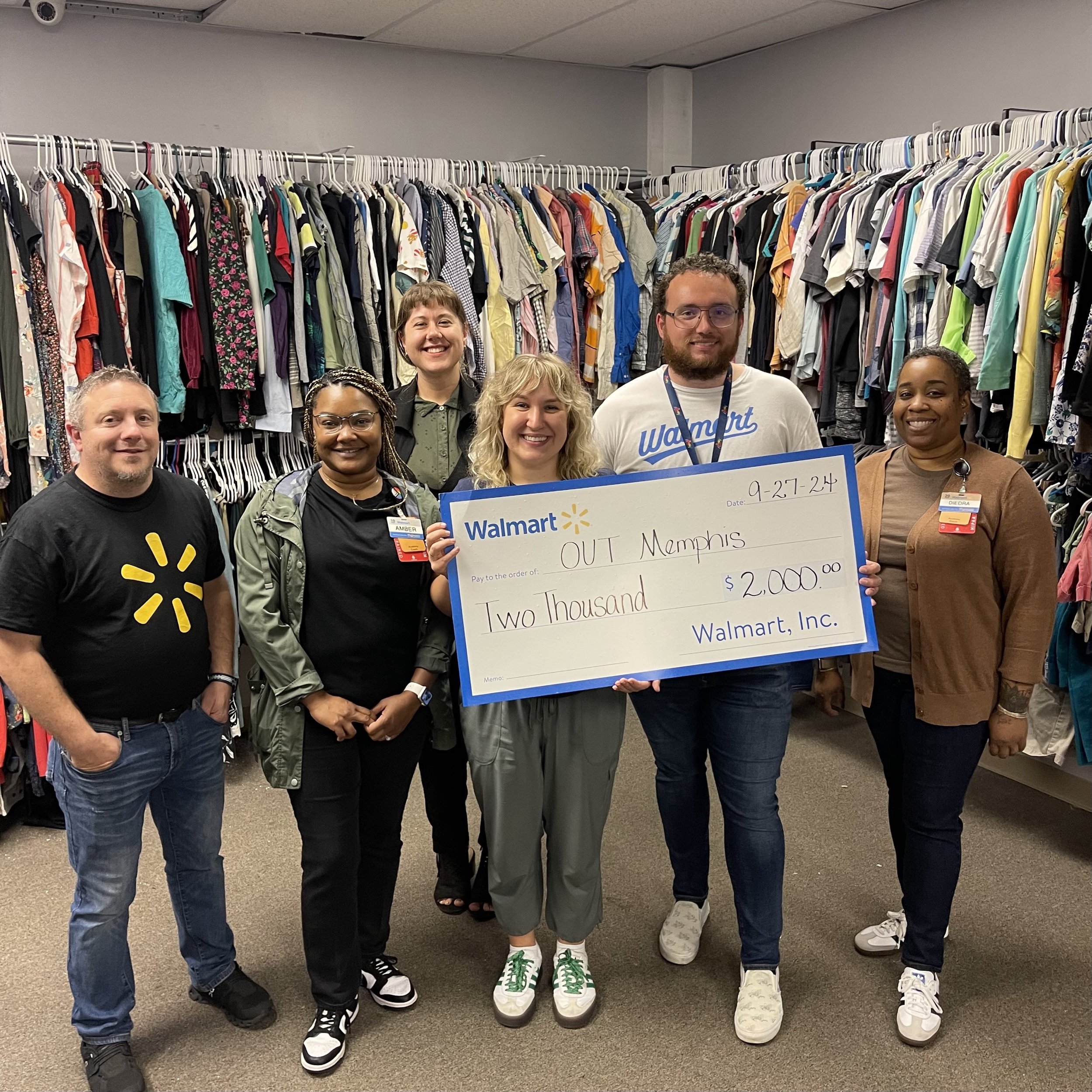 Group of six people holding a large check from Walmart for $2,000 made out to OUTMemphis, standing in front of racks of clothes. The check is dated 9-27-24. Some individuals are wearing Walmart attire with name badges.