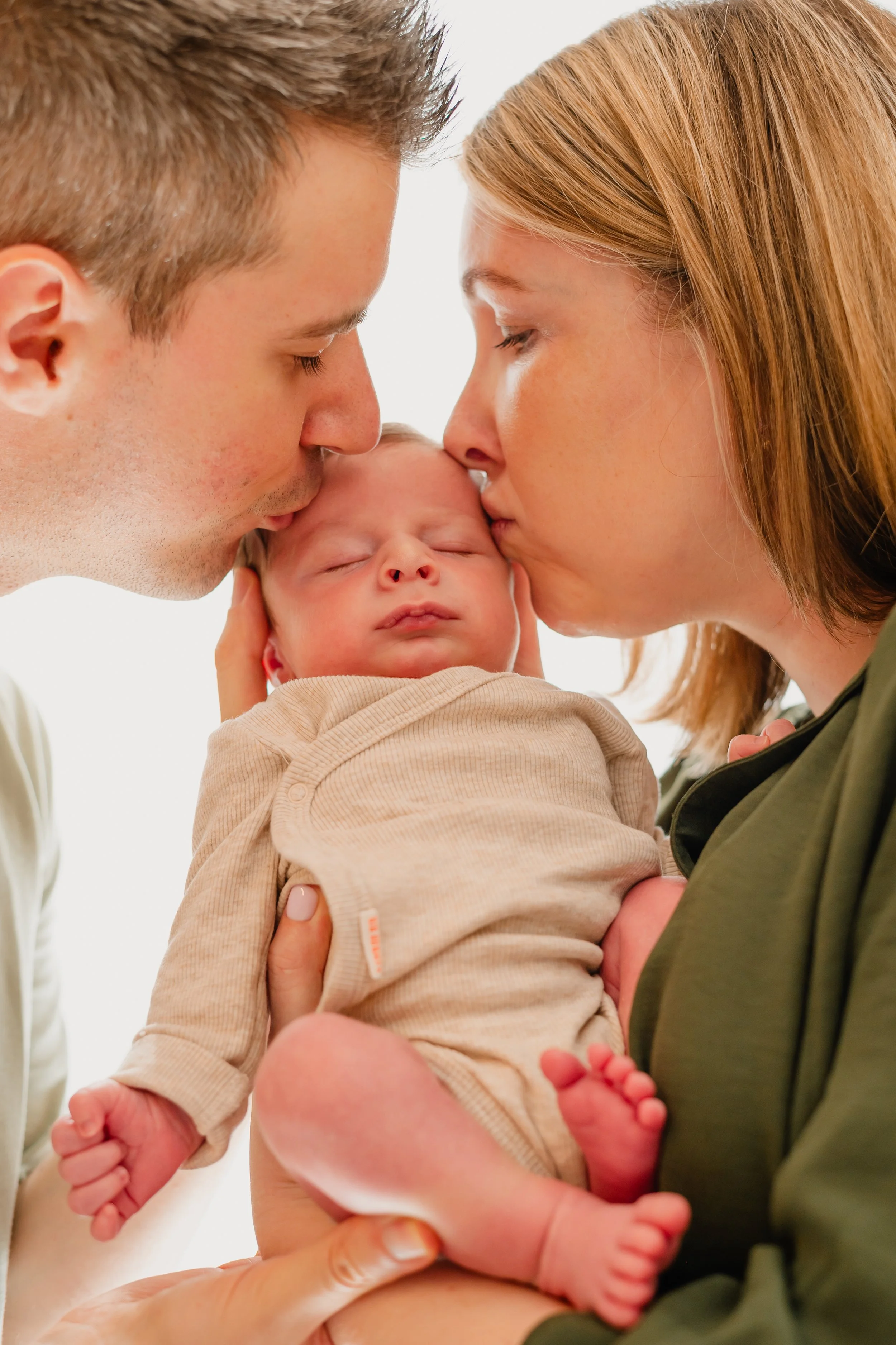 Mama en papa kussen een slapende baby op het hoofd en wang, met witte achtergrond.