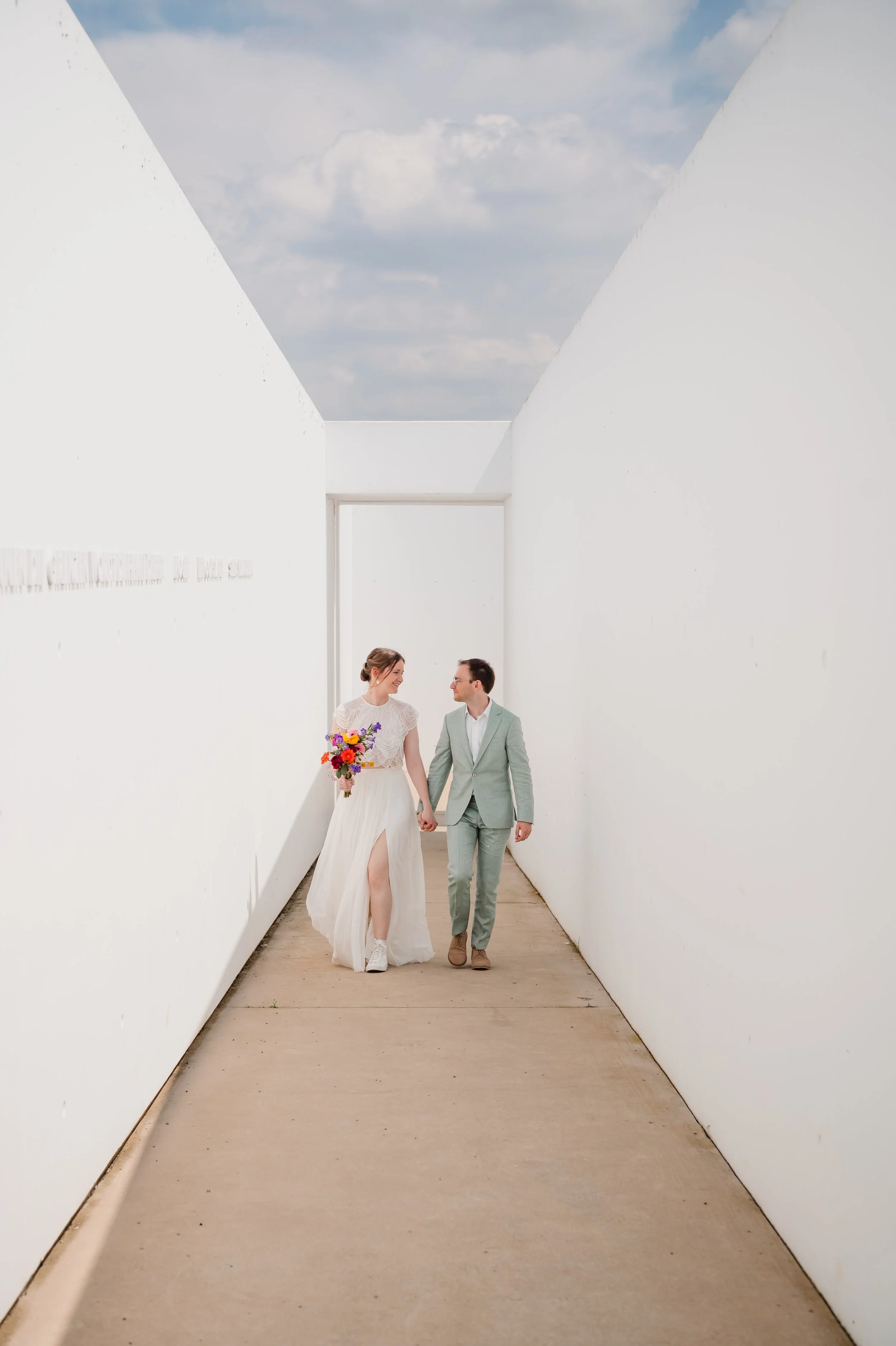 Trouwpaar wandelend hand in hand door een witte, minimalistische gang onder een blauwe hemel. Rumbeke.