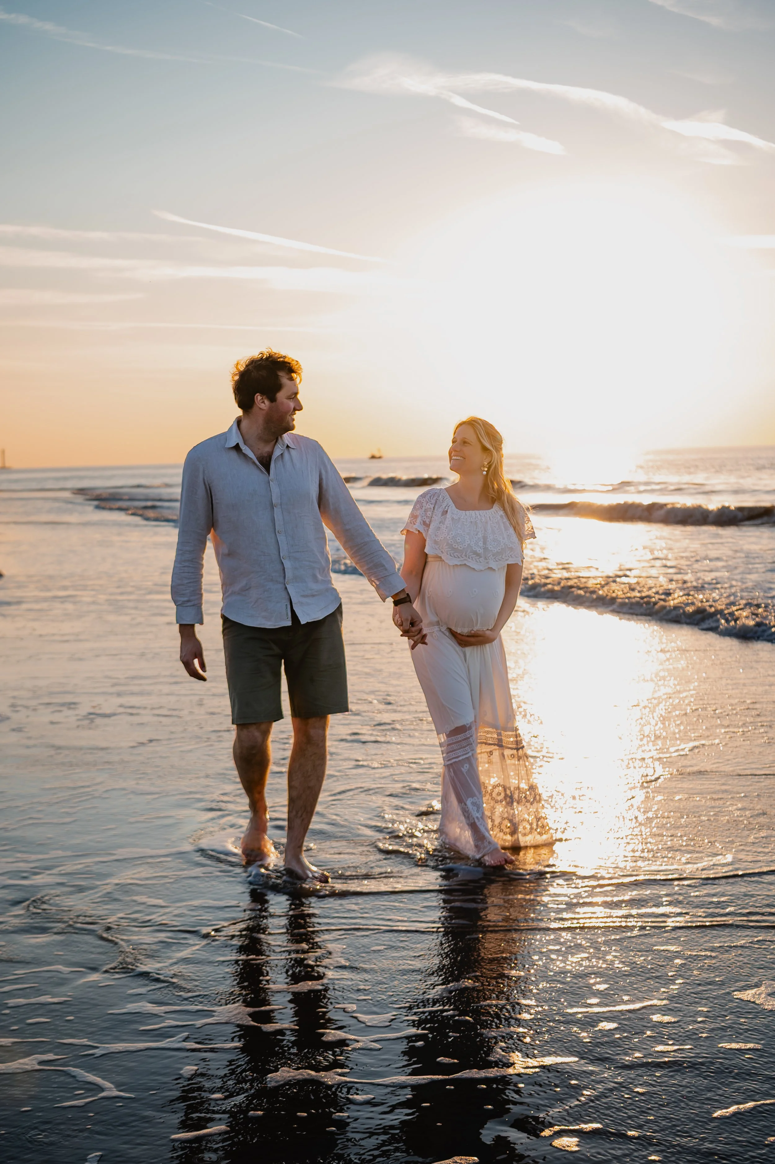 Een zwangere vrouw en haar partner lopen hand in hand op het strand bij zonsondergang.