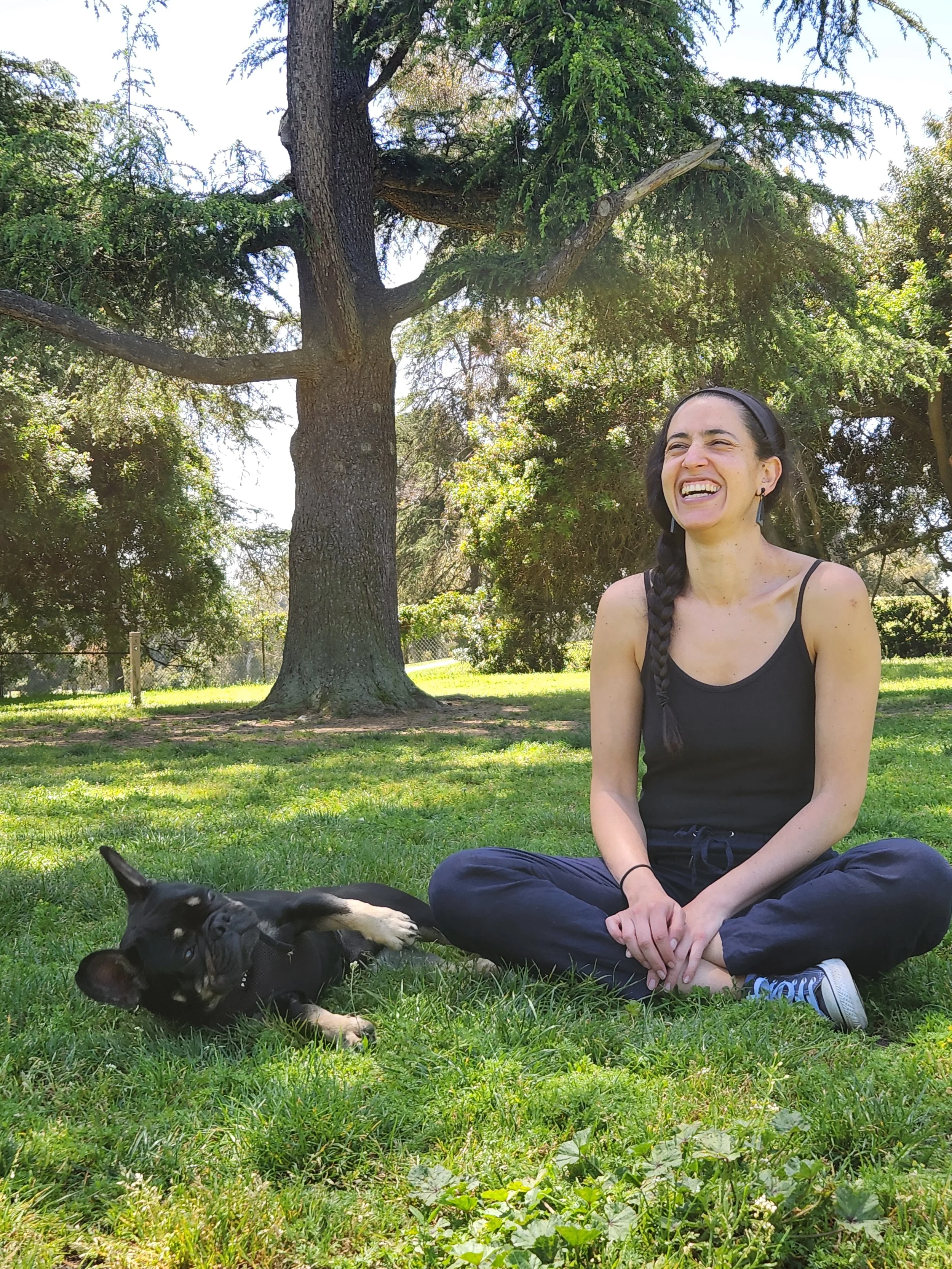 A woman with long dark braided hair, sitting cross-legged on grass in a park, laughing. A black and white puppy is lying beside her, also on the grass. Behind them are trees with green leaves and a sunny sky.