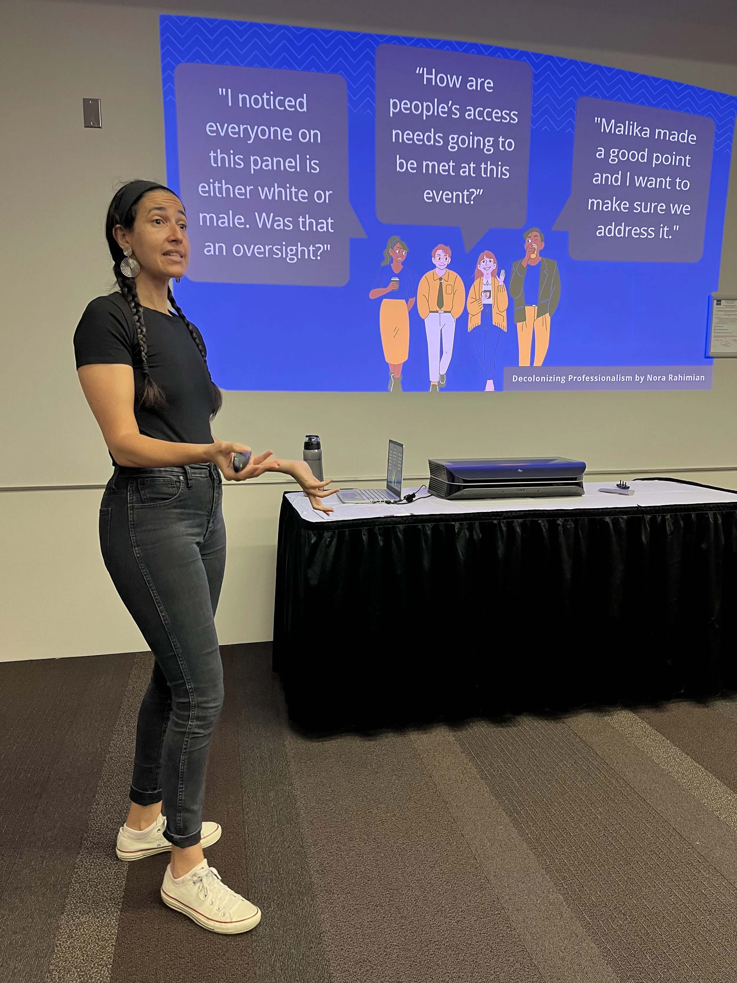 A woman standing in front of a presentation slide, gesturing with her hands. She has long braided hair, is wearing black jeans, a black t-shirt, and white sneakers. The slide behind her shows illustrations of people with speech bubbles containing tex