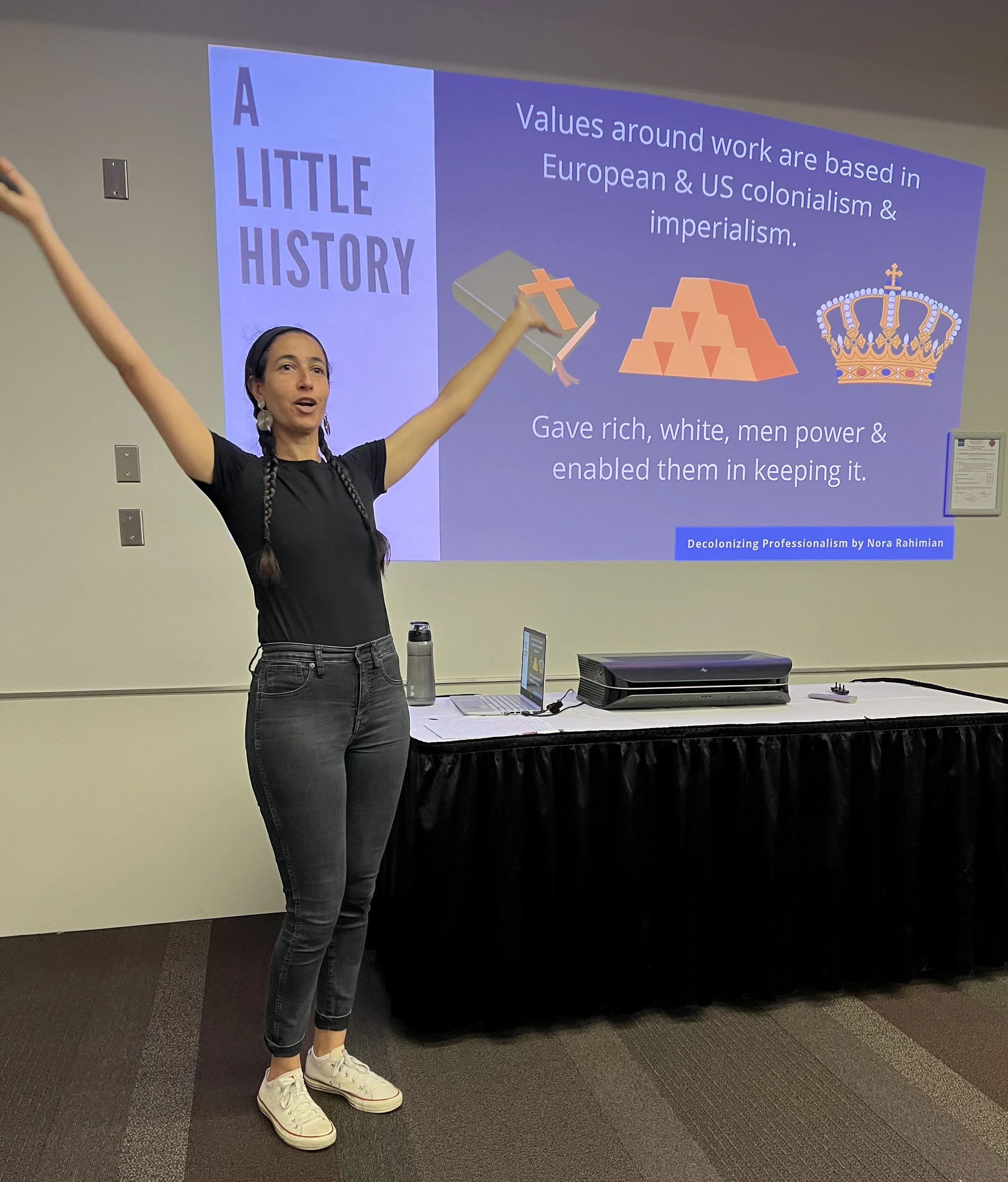 A woman with braided hair, wearing a black shirt, dark jeans, and white sneakers, standing in front of a presentation screen with her arms raised. The presentation slide behind her has the title 'A Little History' and includes a graphic of a Bible with a cross, a pyramid with triangles, and a royal crown. The slide also contains text about colonialism, imperialism, and the history of European and US influence.