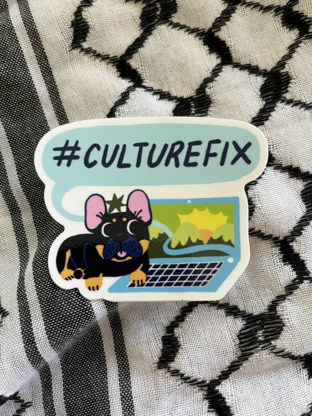 A sticker with the hashtag #CULTUREFIX, featuring a cartoon pig wearing a crown, with a laptop showing a landscape with mountains and a sun, on a black and white patterned fabric background.