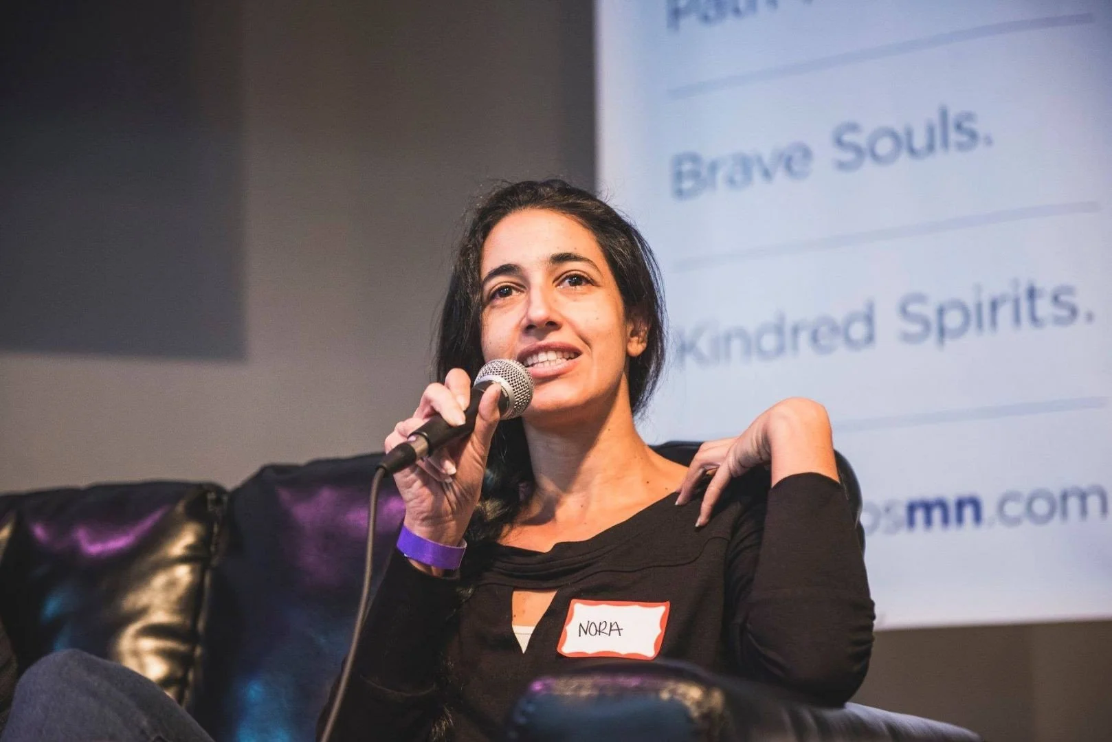 A woman with dark hair holding a microphone and sitting on a black leather chair. She is wearing a black top and has a red and white name tag that says Nora. Behind her is a screen with partially visible blue text.