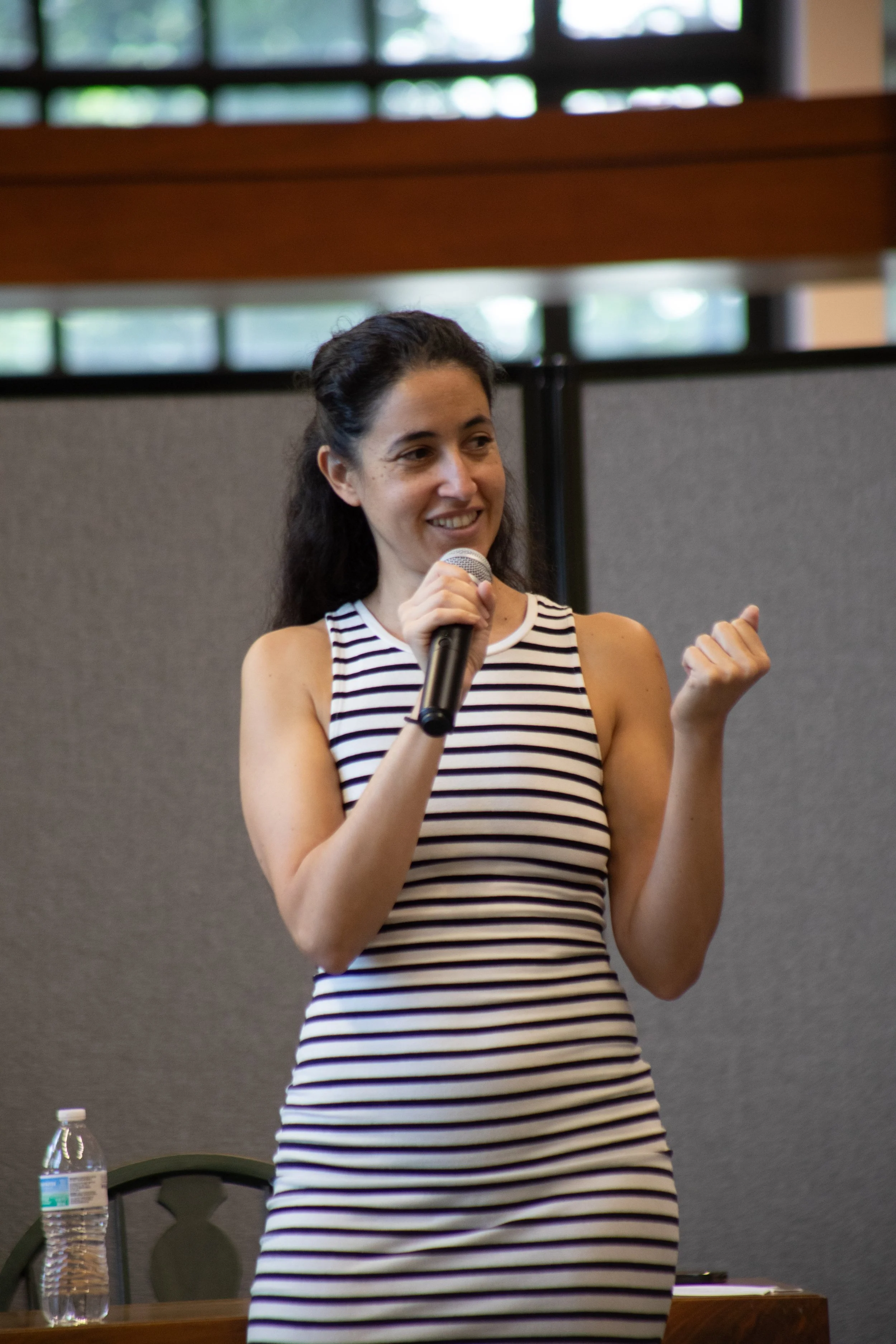 A woman with dark hair wearing a sleeveless, striped top, holding a microphone and gesturing with her right hand, smiling during a presentation or speech. There is a water bottle on the table in front of her, and a gray partition behind her.