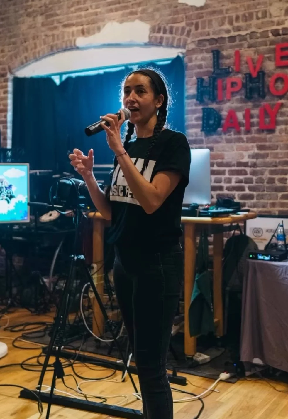 A woman with braided hair singing into a microphone at an event with a brick wall background and red and blue 'LIVE HIPHOP DAY' sign, surrounded by DJ equipment and wires.
