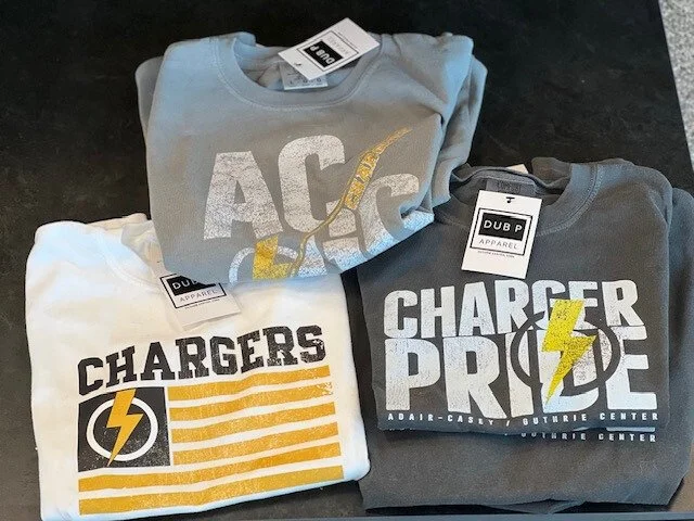 Three folded t-shirts with Chargers Pride and Charger logos, on a black surface.