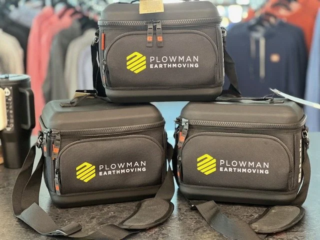 Three black carrying cases with zippers and handles, labeled 'Plowman Earthmoving' with a yellow and black logo, on a table in a store.