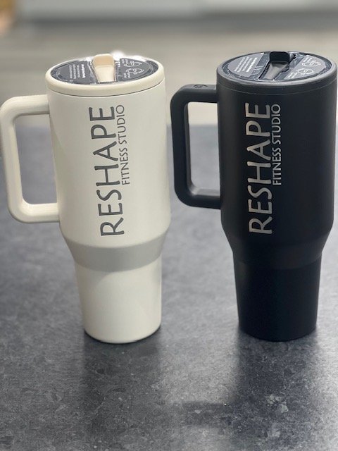 White and black ReShape Fitness Studio travel mugs with lids on a dark surface.