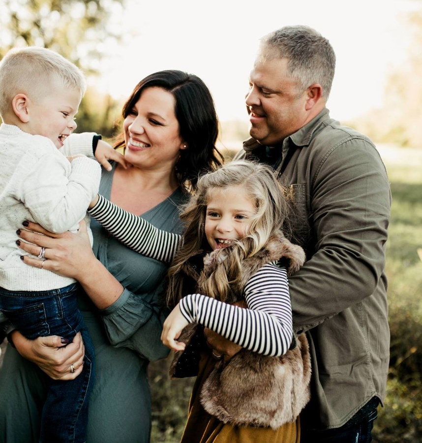 A happy family of four outdoors, with the parents holding their children, all smiling and enjoying a sunny day.