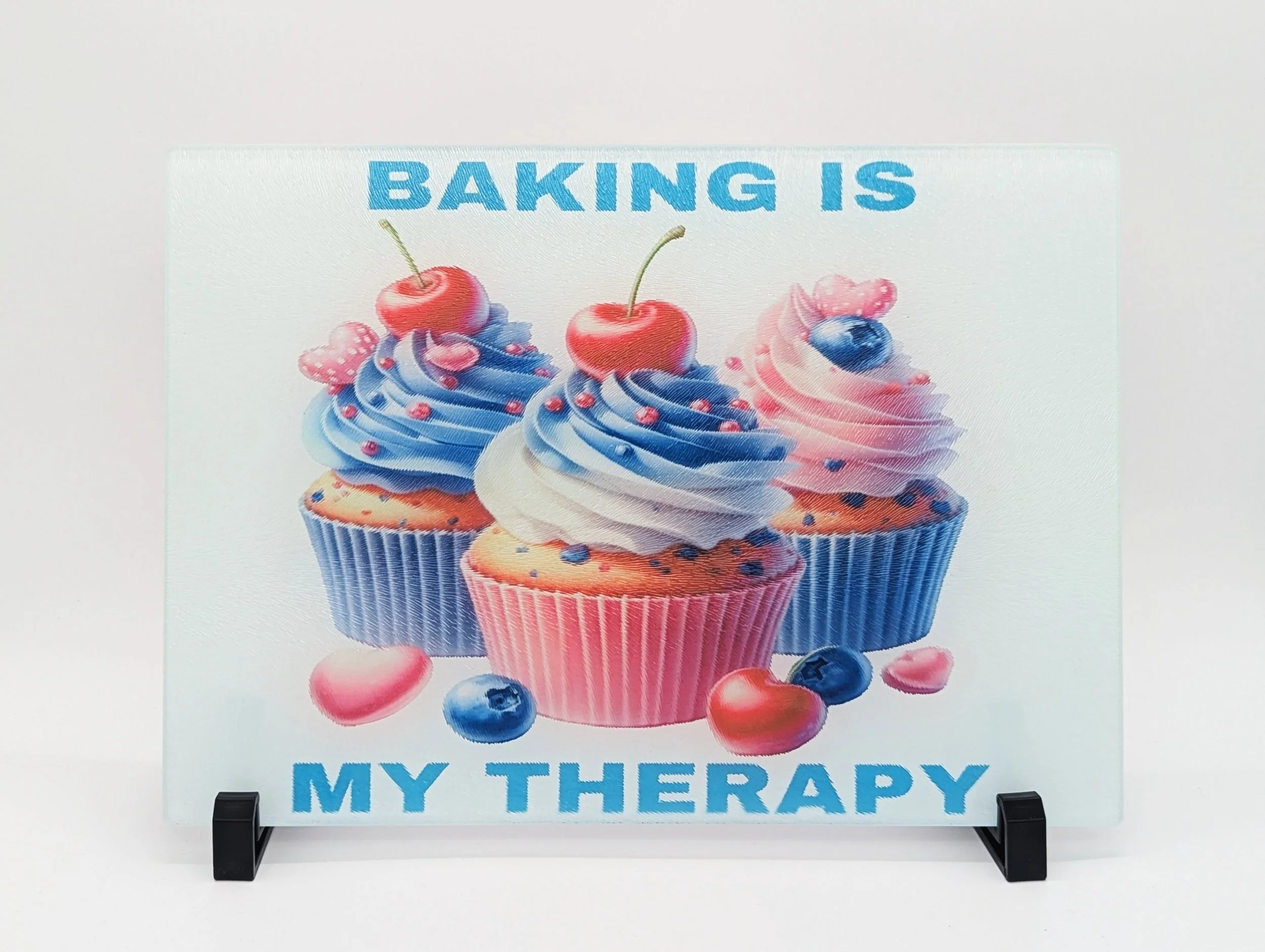 A decorative sign with images of three cupcakes topped with blue, white, and pink swirled frosting, decorated with cherries, blueberries, and pink heart-shaped candies. The sign has the text "Baking is my therapy" in bold blue letters.