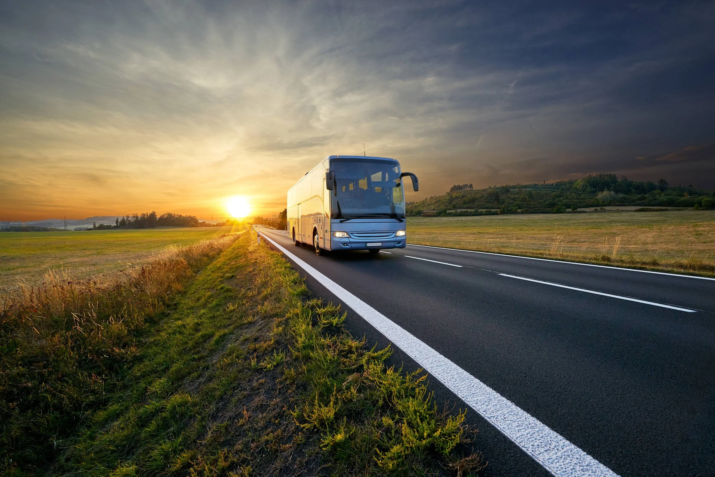 Refinished motor coach bus driving down a rural highway at sunset, surrounded by open fields and rolling countryside beneath a dramatic evening sky.