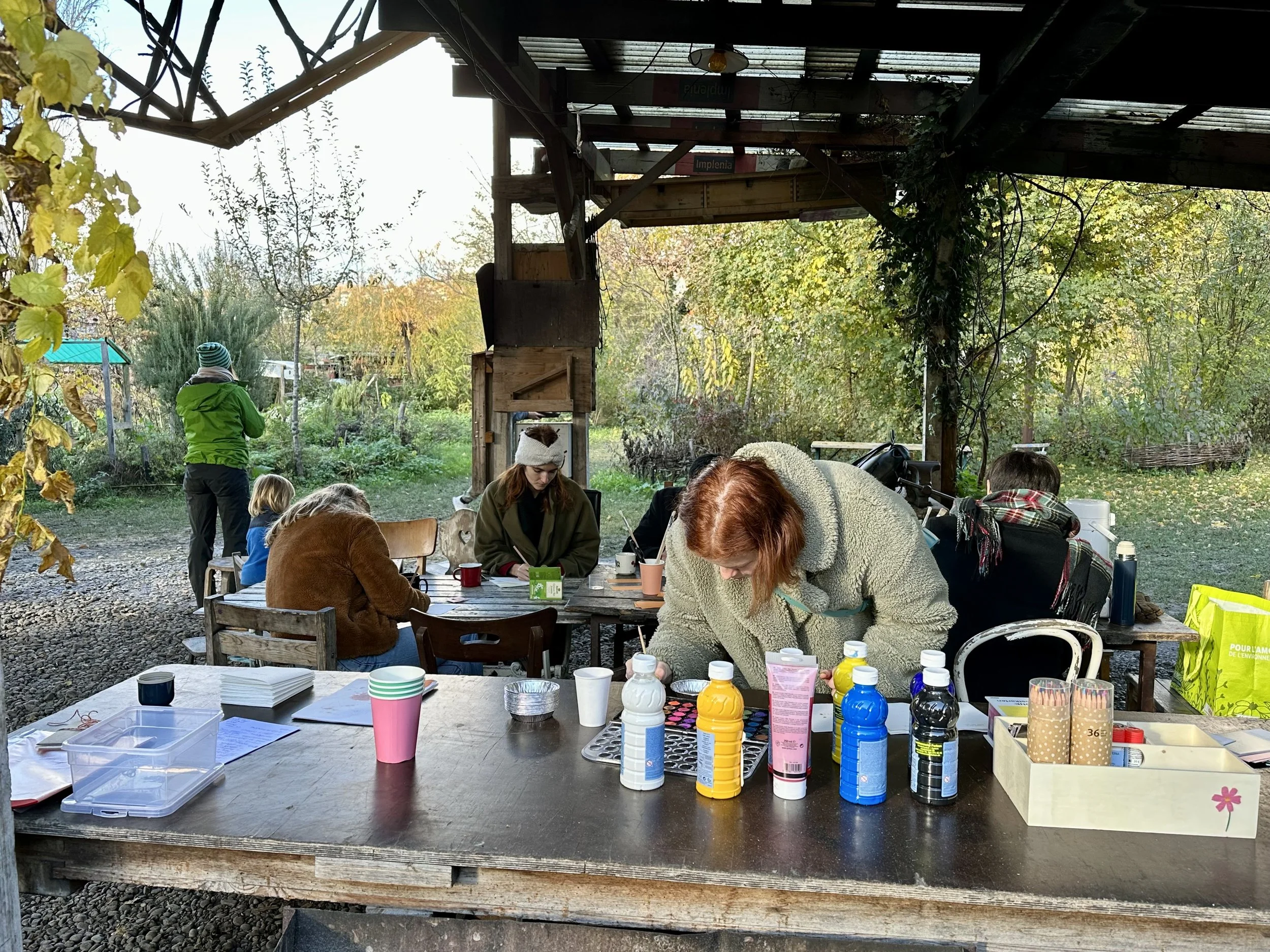People gathered around outdoor table engaging in arts and crafts, with various bottles, cups, and supplies on the table, surrounded by trees and greenery.
