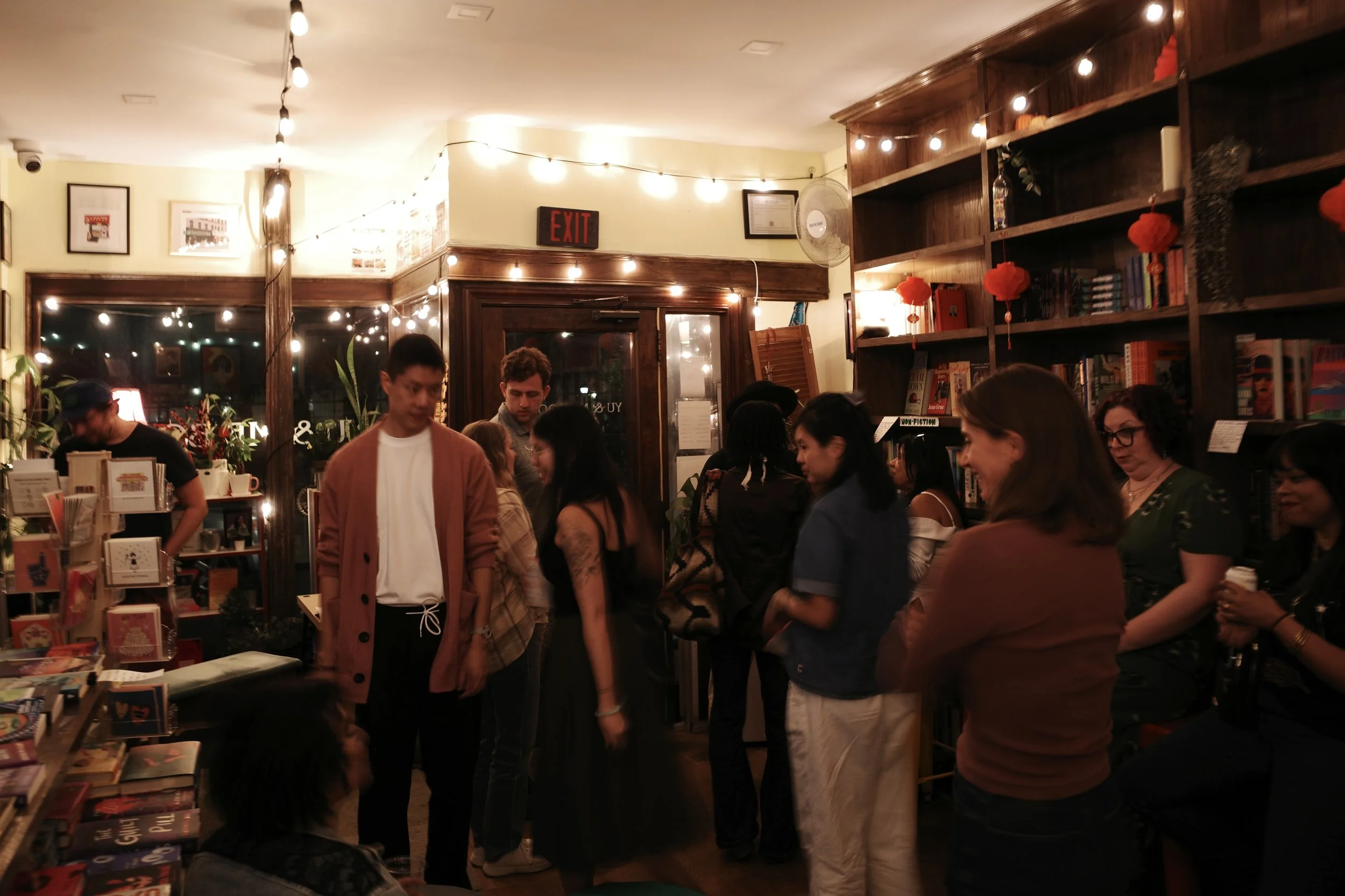 People inside a bookstore or library with shelves of books, string lights hanging from the ceiling, and some individuals browsing or waiting in line.