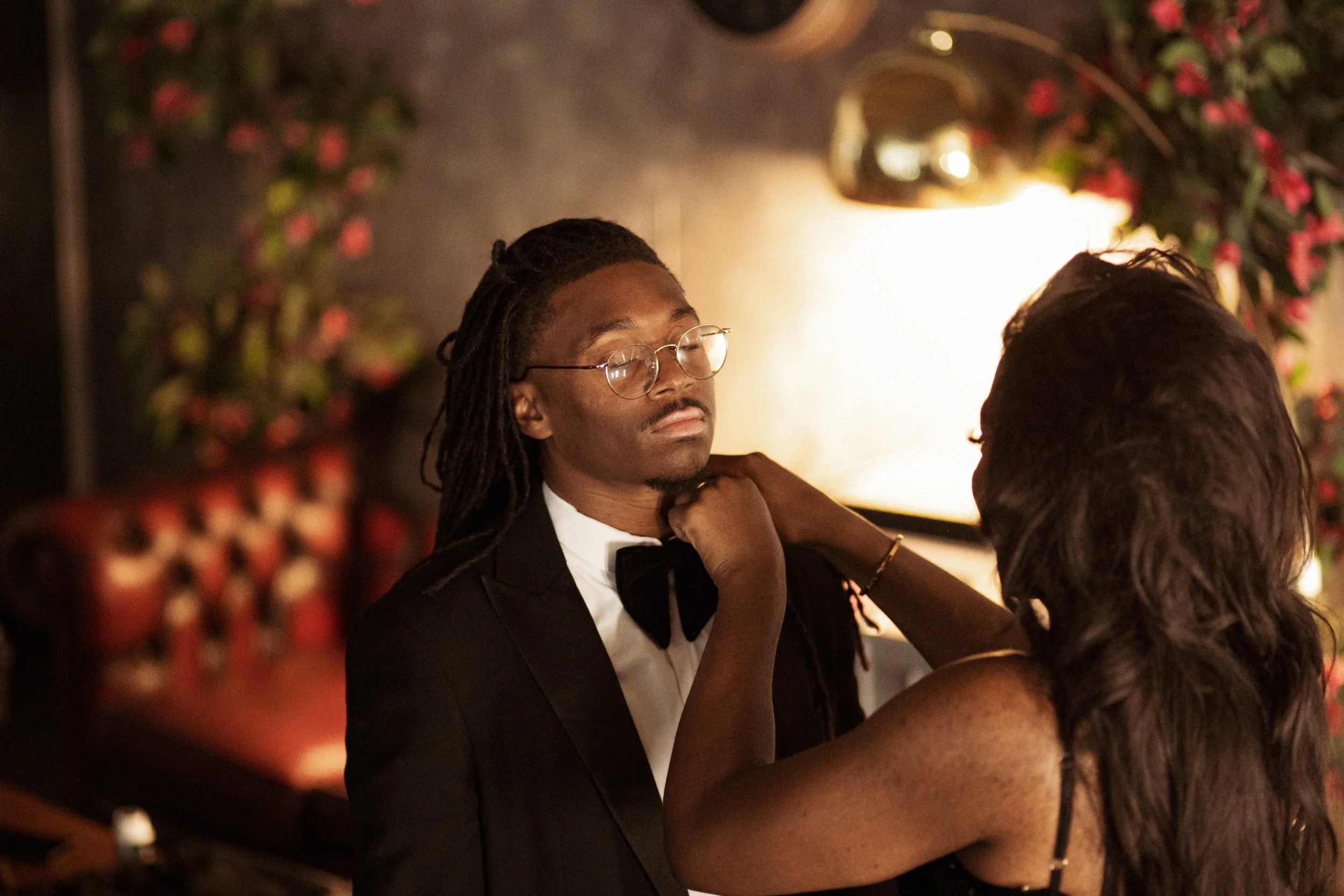 A woman adjusts a man's bow tie in a dimly lit setting with floral decorations and warm lighting.