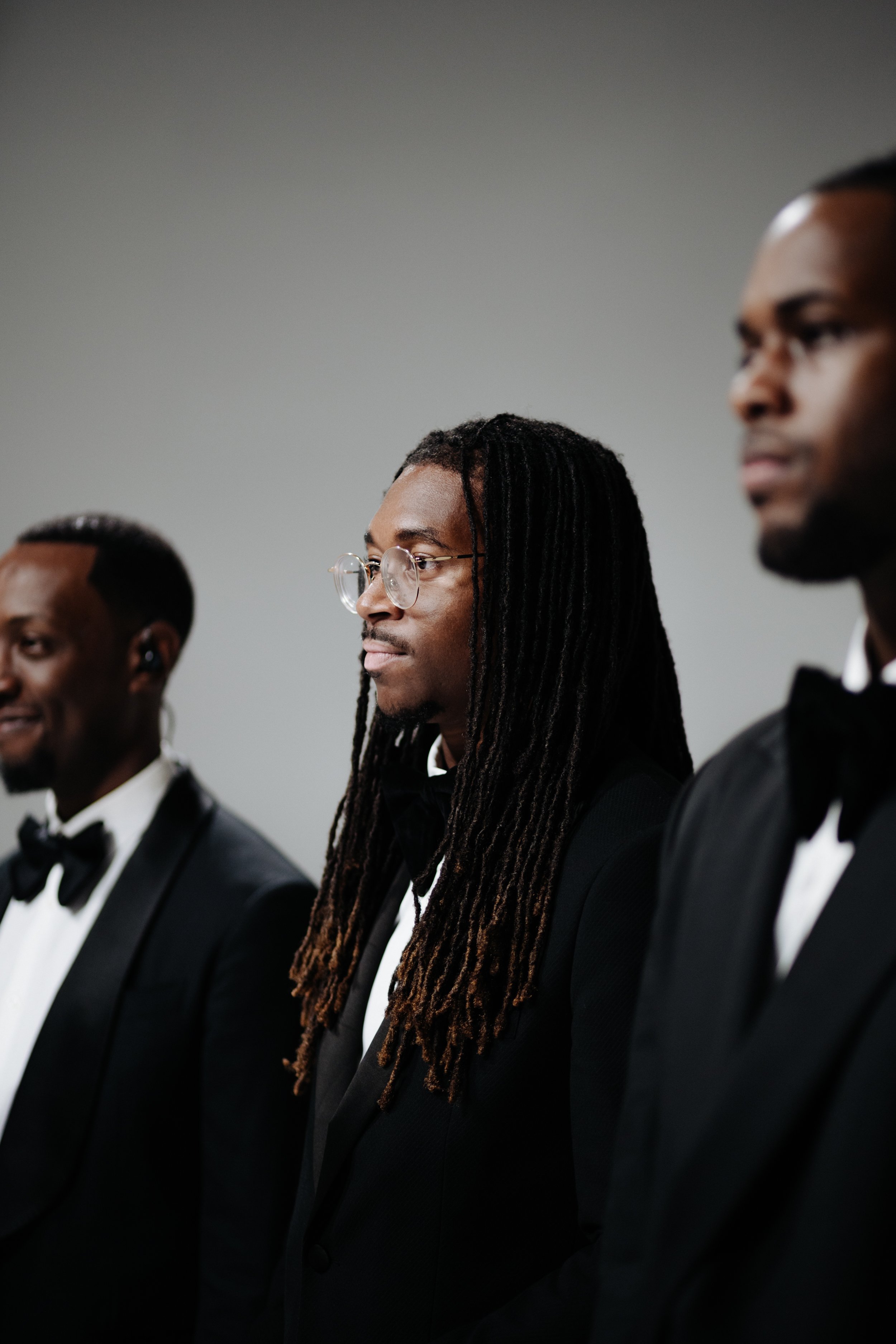 Three men dressed in black tuxedos with bow ties, standing in a row against a plain background. The man in the middle has long dreadlocks and glasses, and appears to be focused or serious. The other two men are smiling slightly.