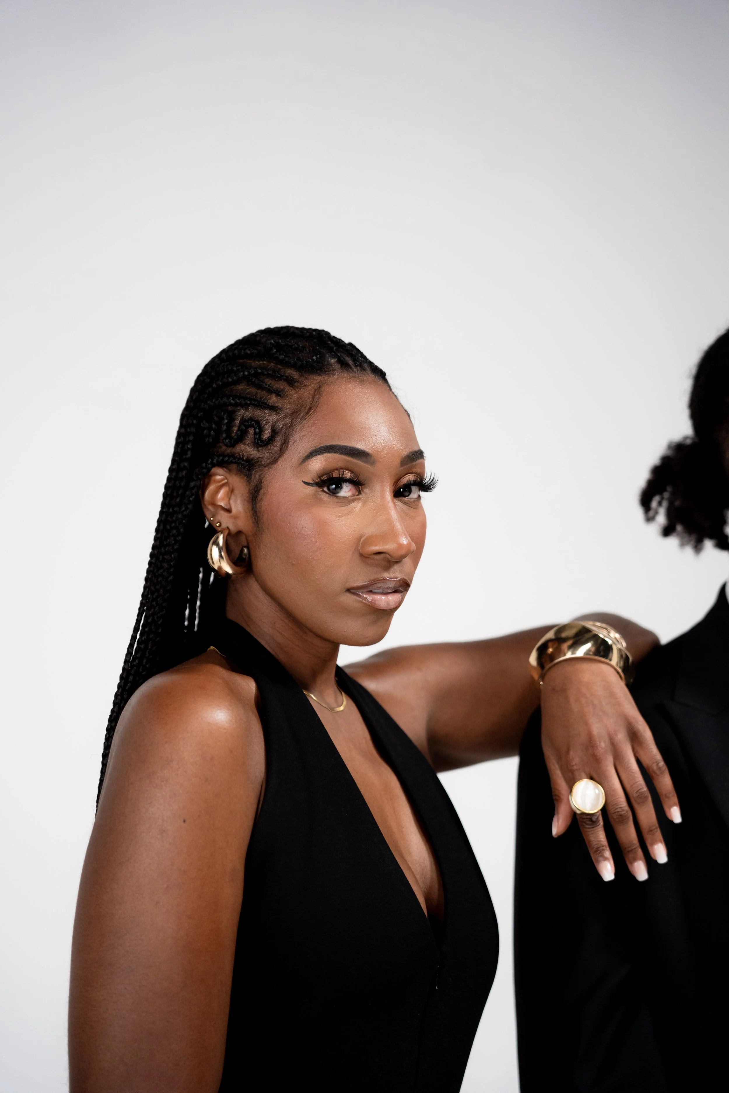 A woman with braided hair and gold jewelry, including earrings, a bracelet, a ring, and a necklace, posing with her arm resting on someone else's shoulder against a plain white background.