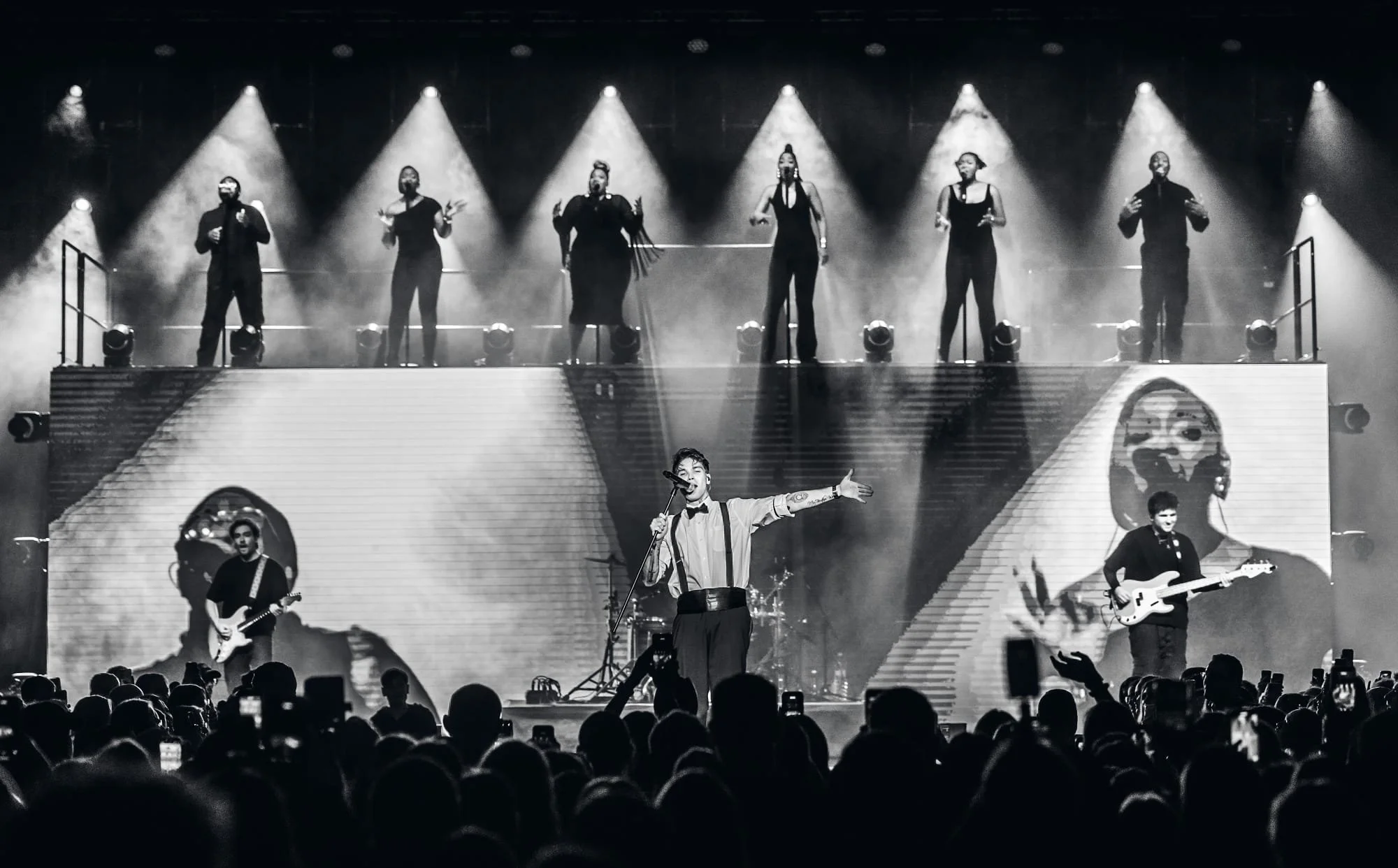 A black and white photo of a live concert with a singer and two guitarists on stage, and six backup singers in the background, with a large screen behind them showing abstract graphics. The audience is visible in the foreground.